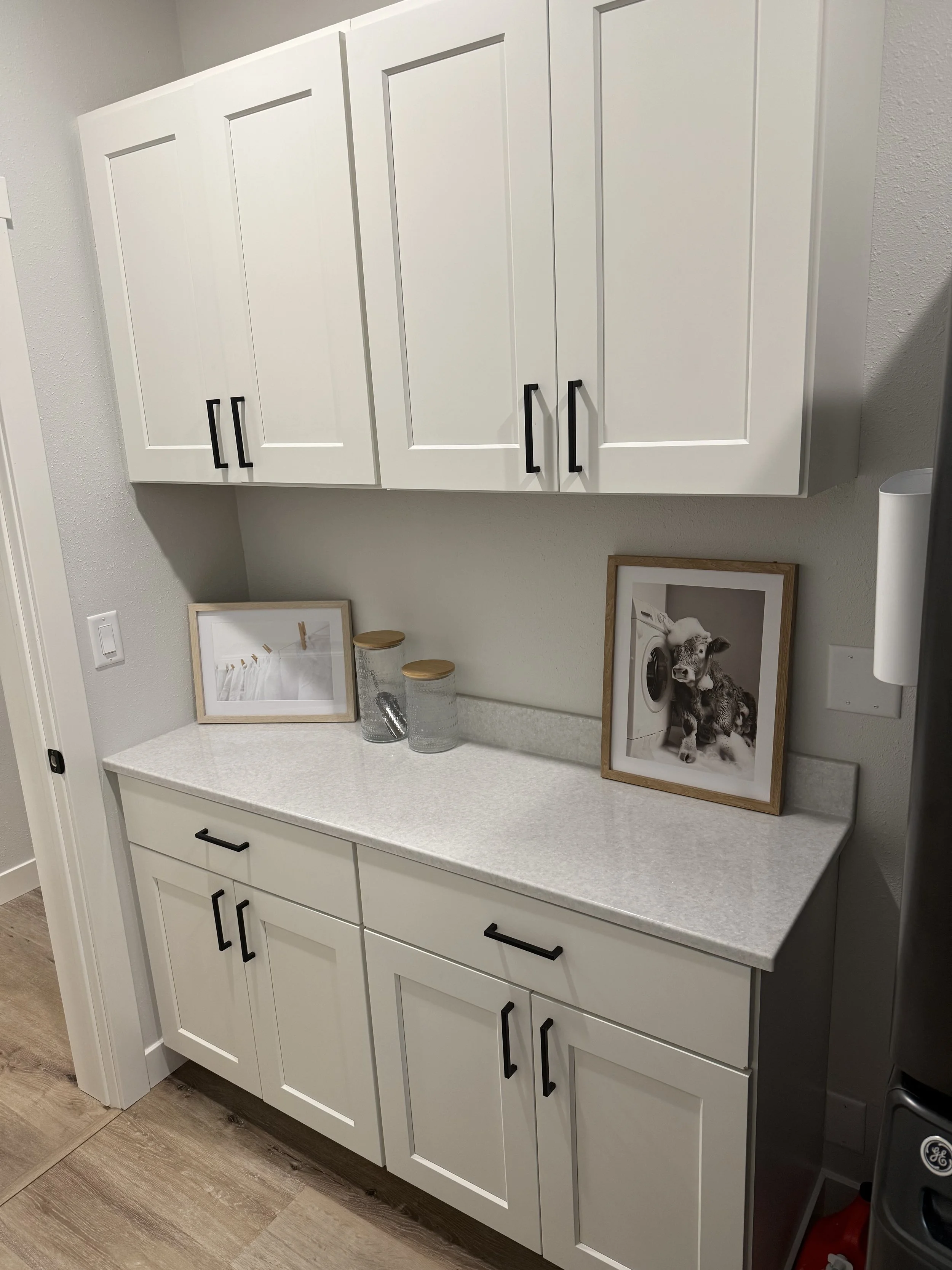 White kitchen cabinet with black handles, countertop with two glass jars, a picture frame, and a framed black and white photo of a goat and a washing machine.