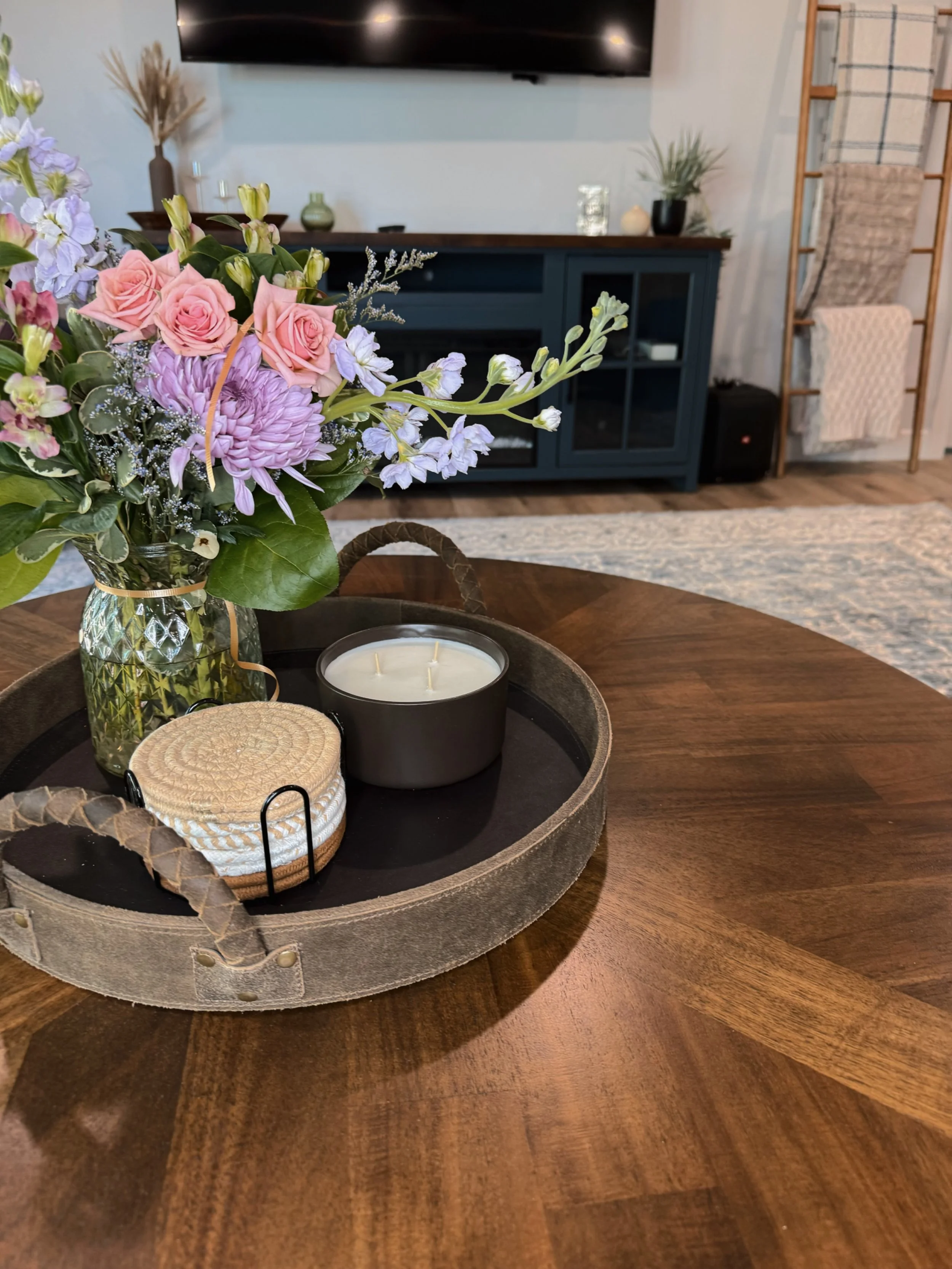 A round wooden table with a black tray holding a vase of pink roses and purple flowers, a lit white candle, and a woven coaster. In the background, a blue TV stand with decorative items and a wall-mounted TV are visible.
