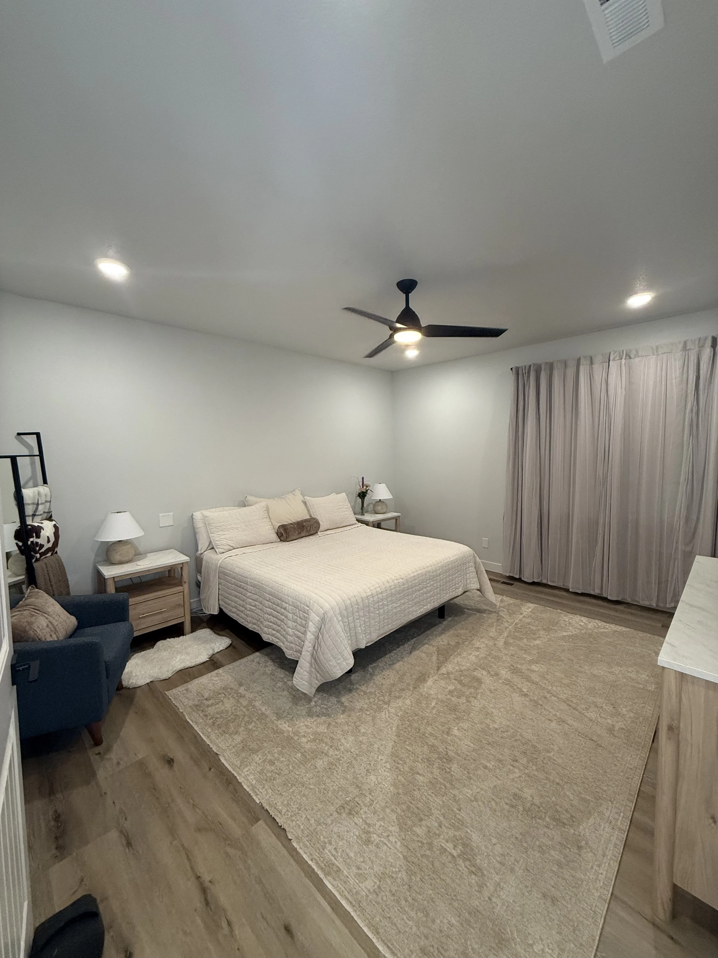 A bedroom with a large bed, beige bedding, and pillows, two nightstands with lamps, a blue armchair, a small rug, and beige curtains covering a window, with white walls and wood flooring.