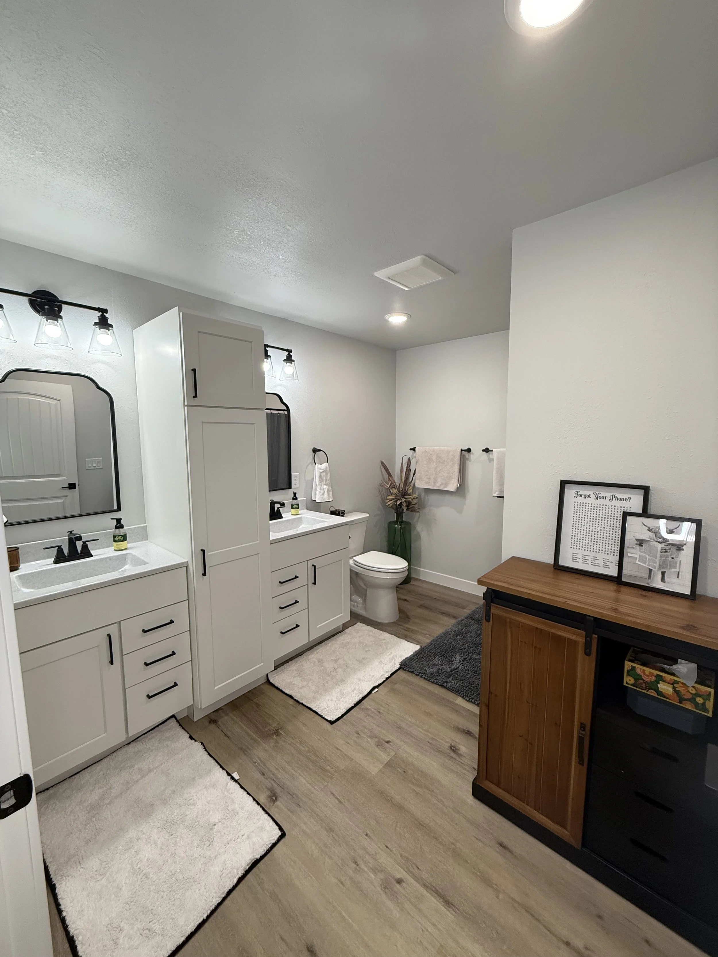 A modern bathroom with dual sinks, mirrors, and vanity lights, a toilet, a tall storage cabinet, and a wooden cabinet with framed pictures, decorated with a plant and towels, on a wood floor with rugs.