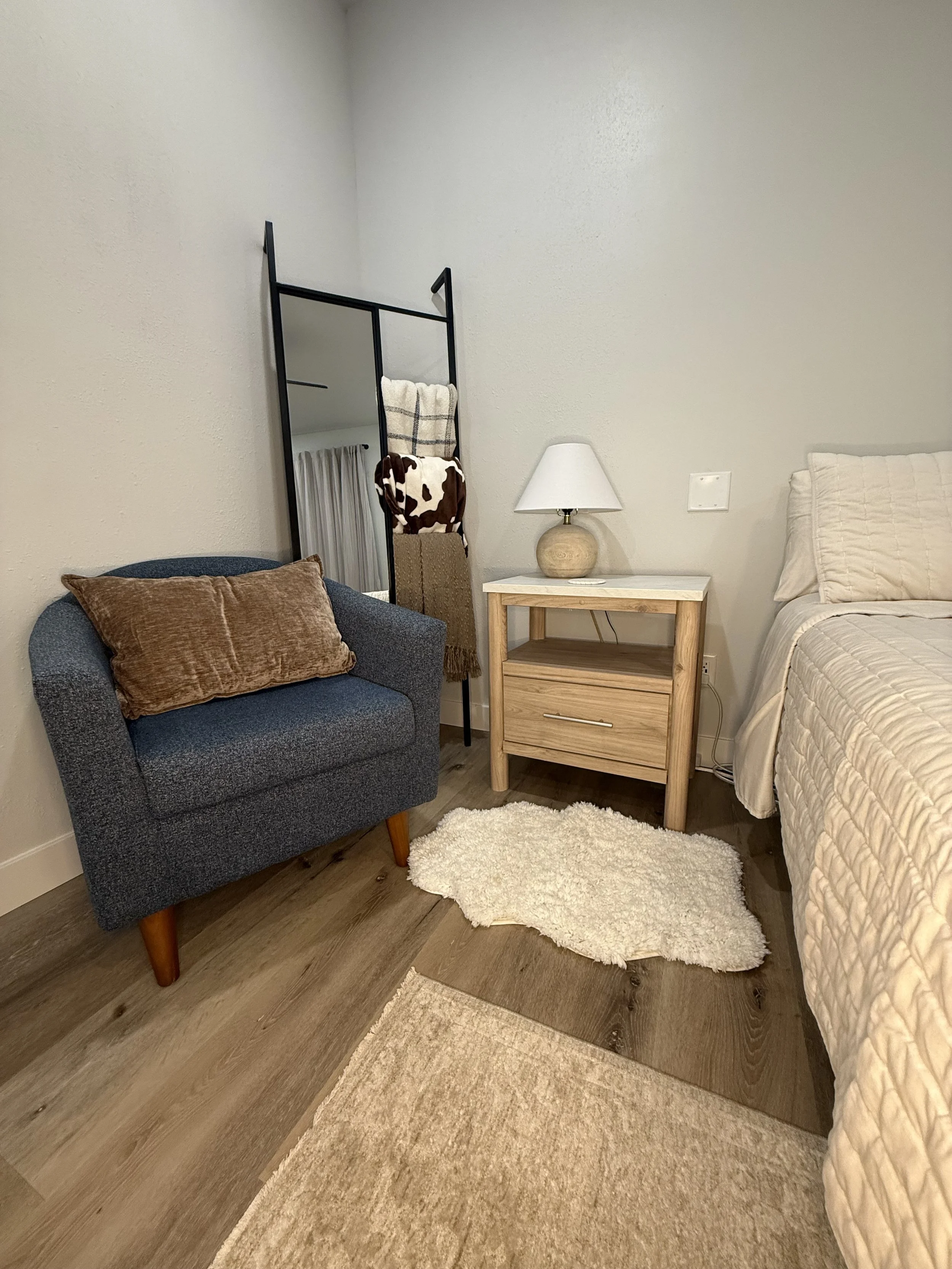 A cozy bedroom corner with a blue armchair, a wooden nightstand with a white lamp, a large bed with cream bedding, a sheepskin rug, and a black metal ladder-style towel rack with blankets hanging on it.