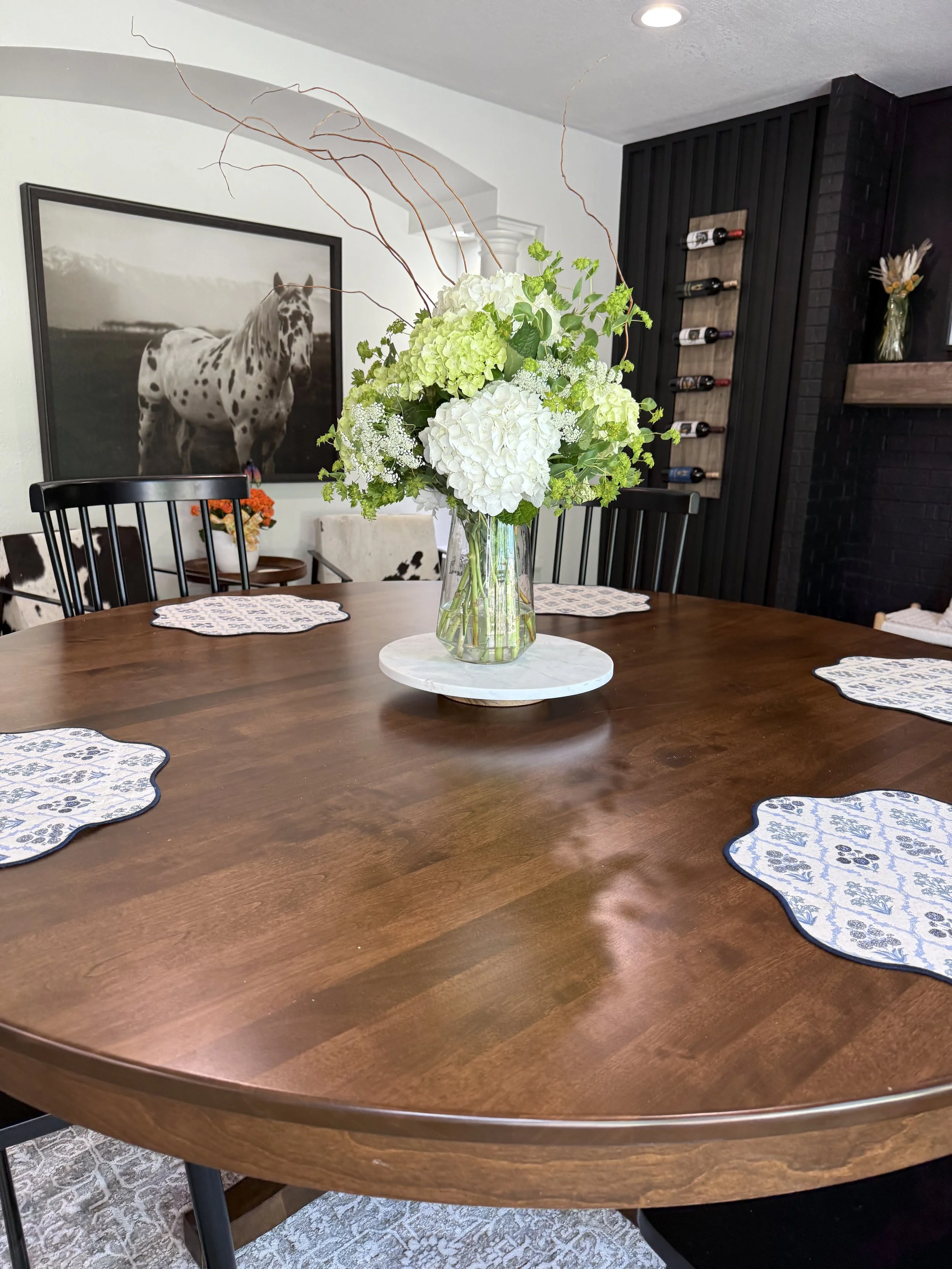 A wooden dining table with a floral centerpiece of white hydrangeas in a glass vase, surrounded by four decorative placemats, in a room with a black and white horse photograph on the wall.