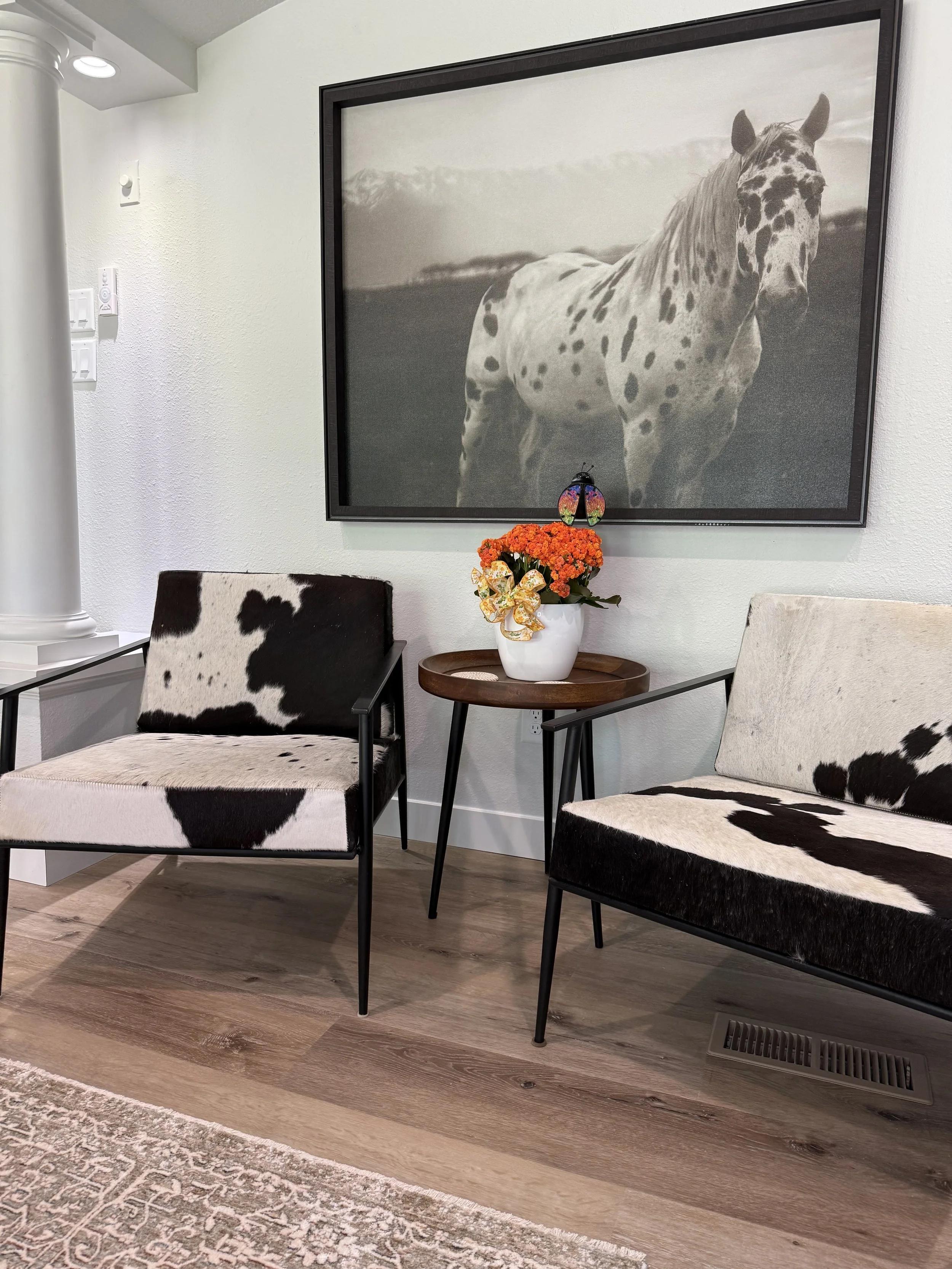 A living room corner with two cowhide-patterned chairs, a small round wooden table with a white vase of orange flowers, and a large black-and-white framed photo of a horse on the wall behind.
