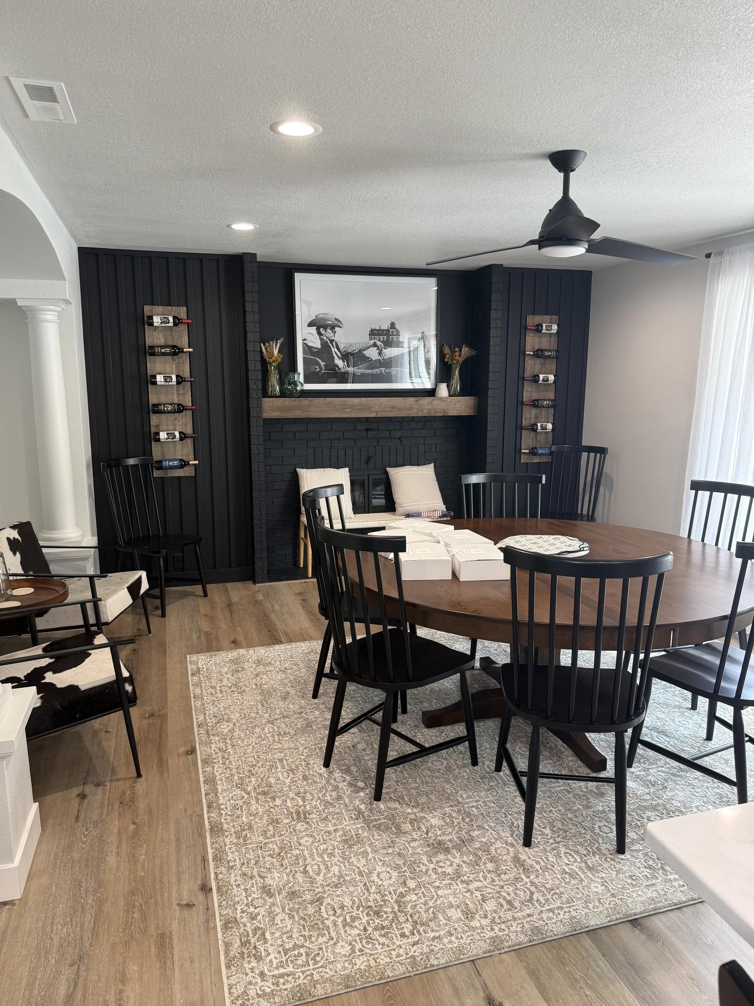 Dining room with a round wooden table surrounded by black chairs, a fireplace with a black brick wall and framed black-and-white photo, wine bottles on wall shelves, and a beige rug on wooden floor.