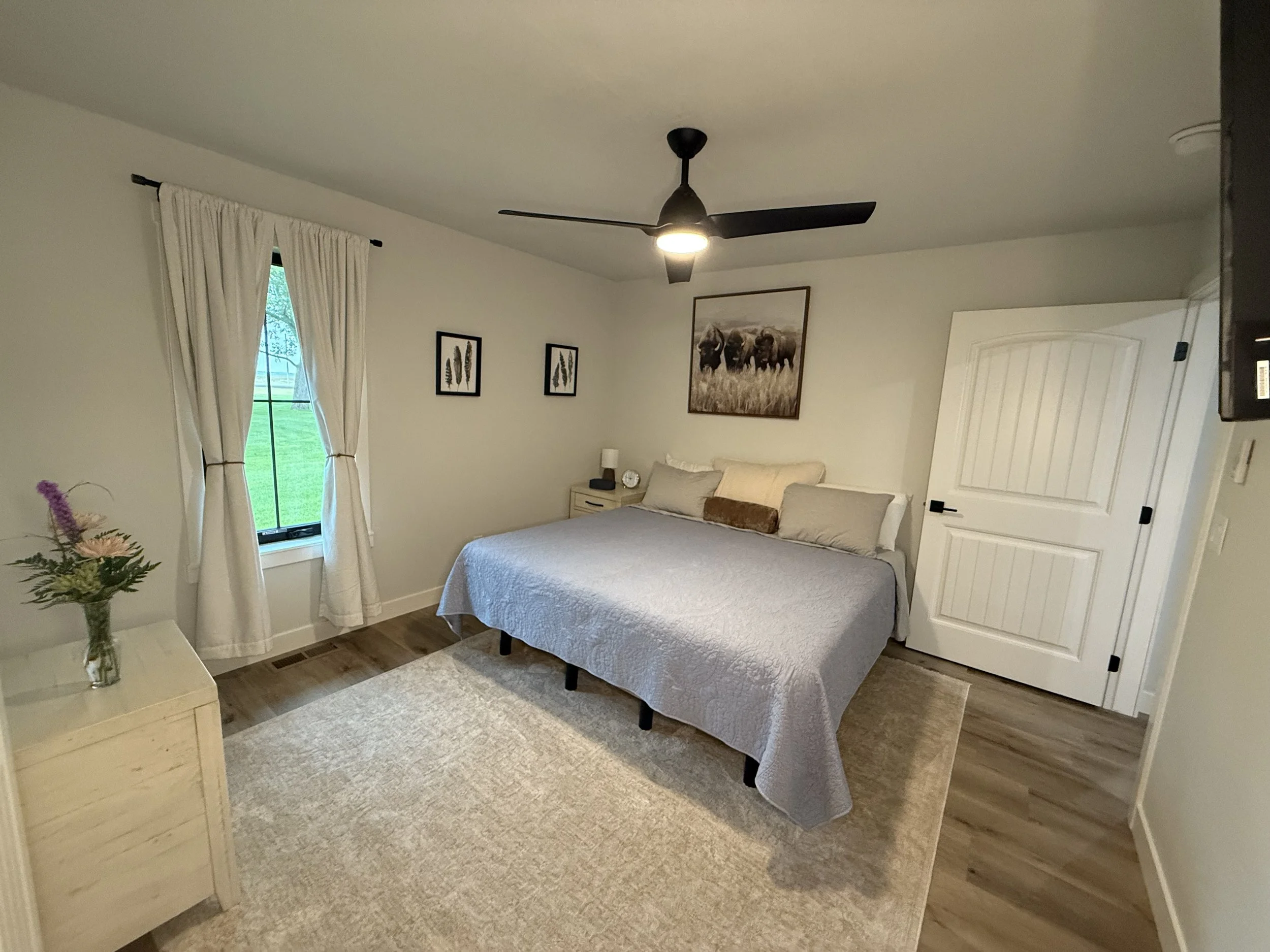 A bedroom with a large bed, a nightstand, wall art, a ceiling fan, a window with white curtains, and a beige rug.