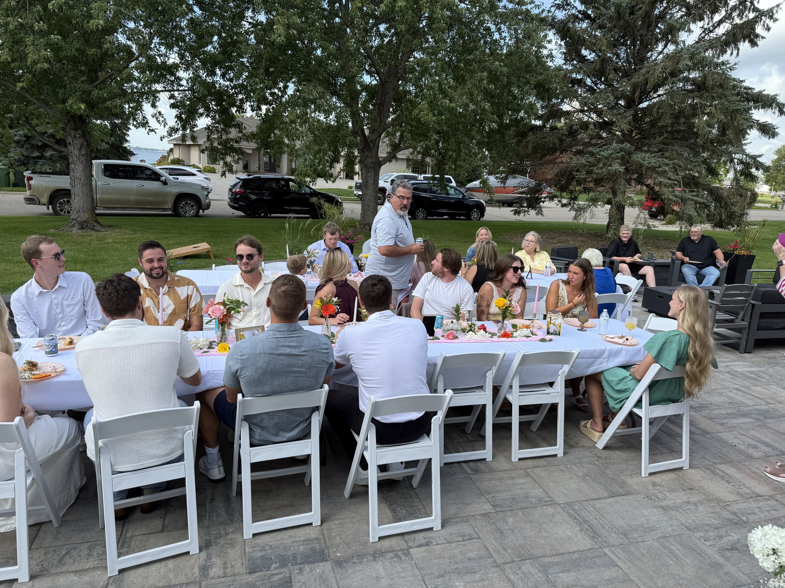 People gathered outdoors at a celebration, seated around a long table with floral centerpieces, with trees and parked cars in the background.