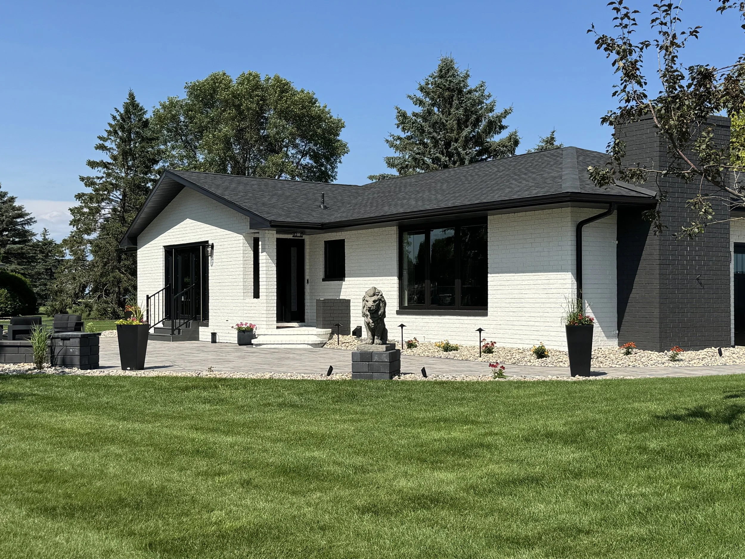 Modern house with black and white exterior, large windows, gray patio, potted plants, a lion statue, and well-maintained lawn under a blue sky.