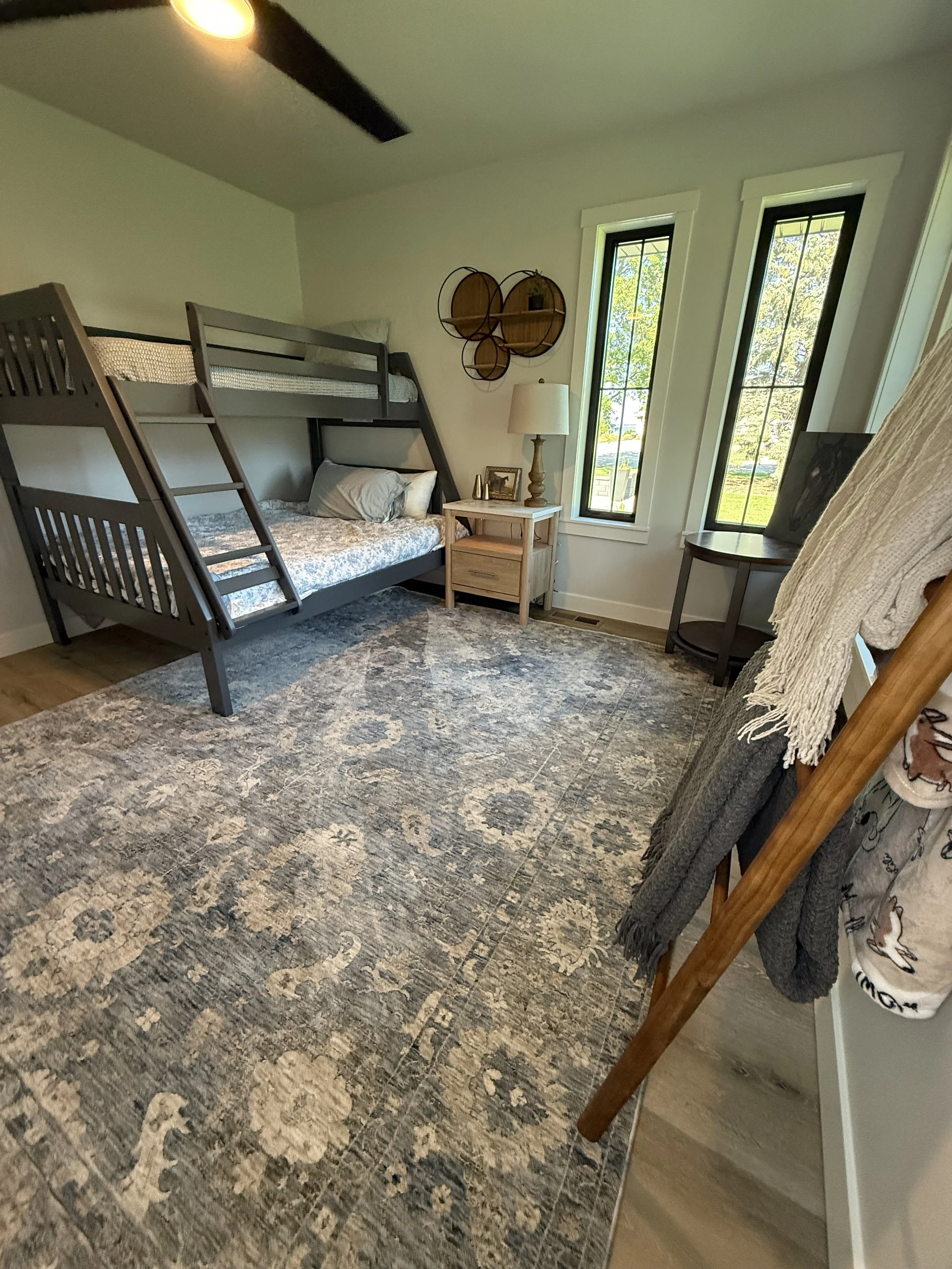 A bedroom with a bunk bed, a side table with a lamp, two windows showing greenery outside, and a cozy rug on the floor.