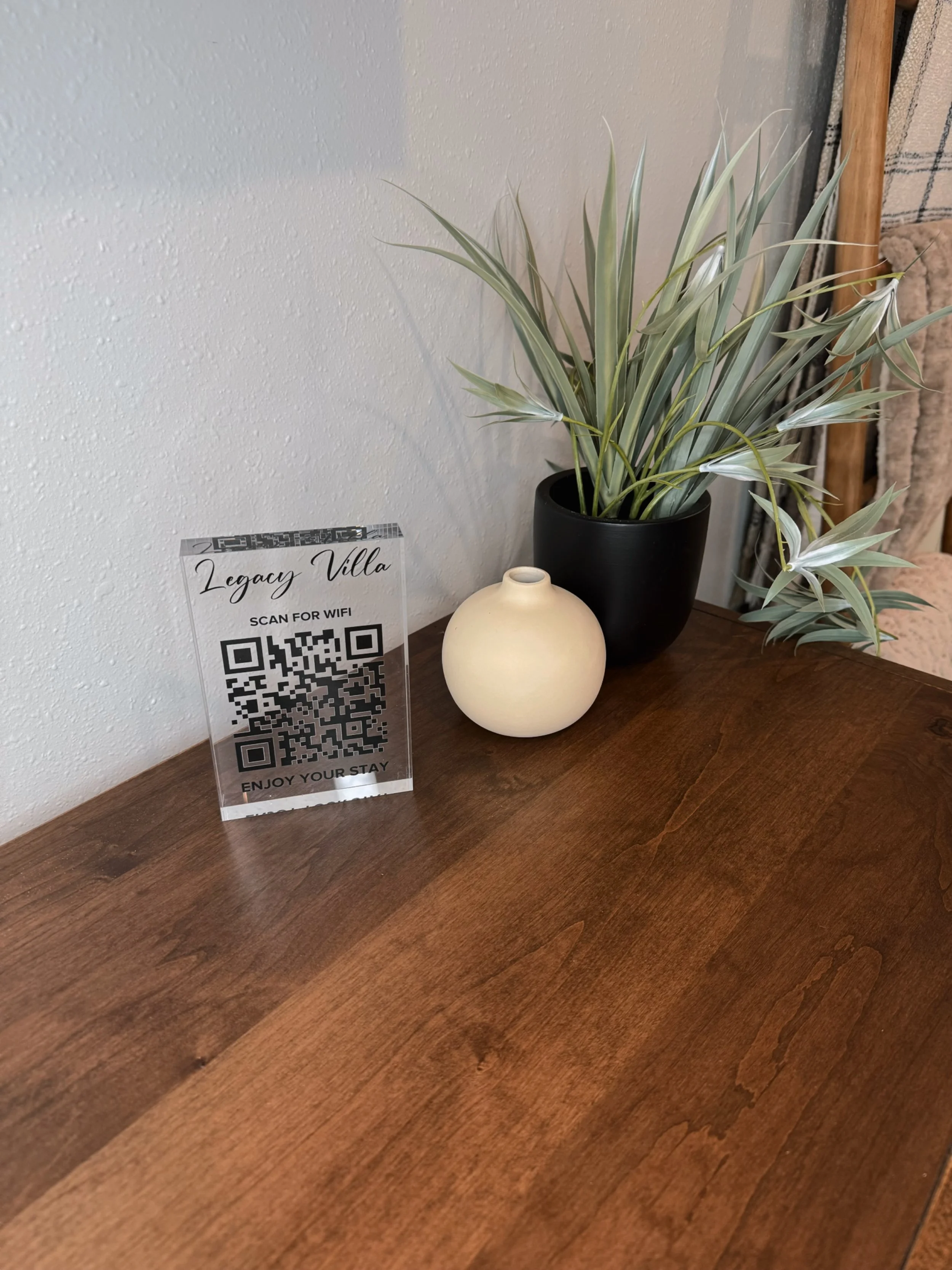 A table with a QR code sign, a black potted plant, and a small ceramic vase in front of a textured gray wall.