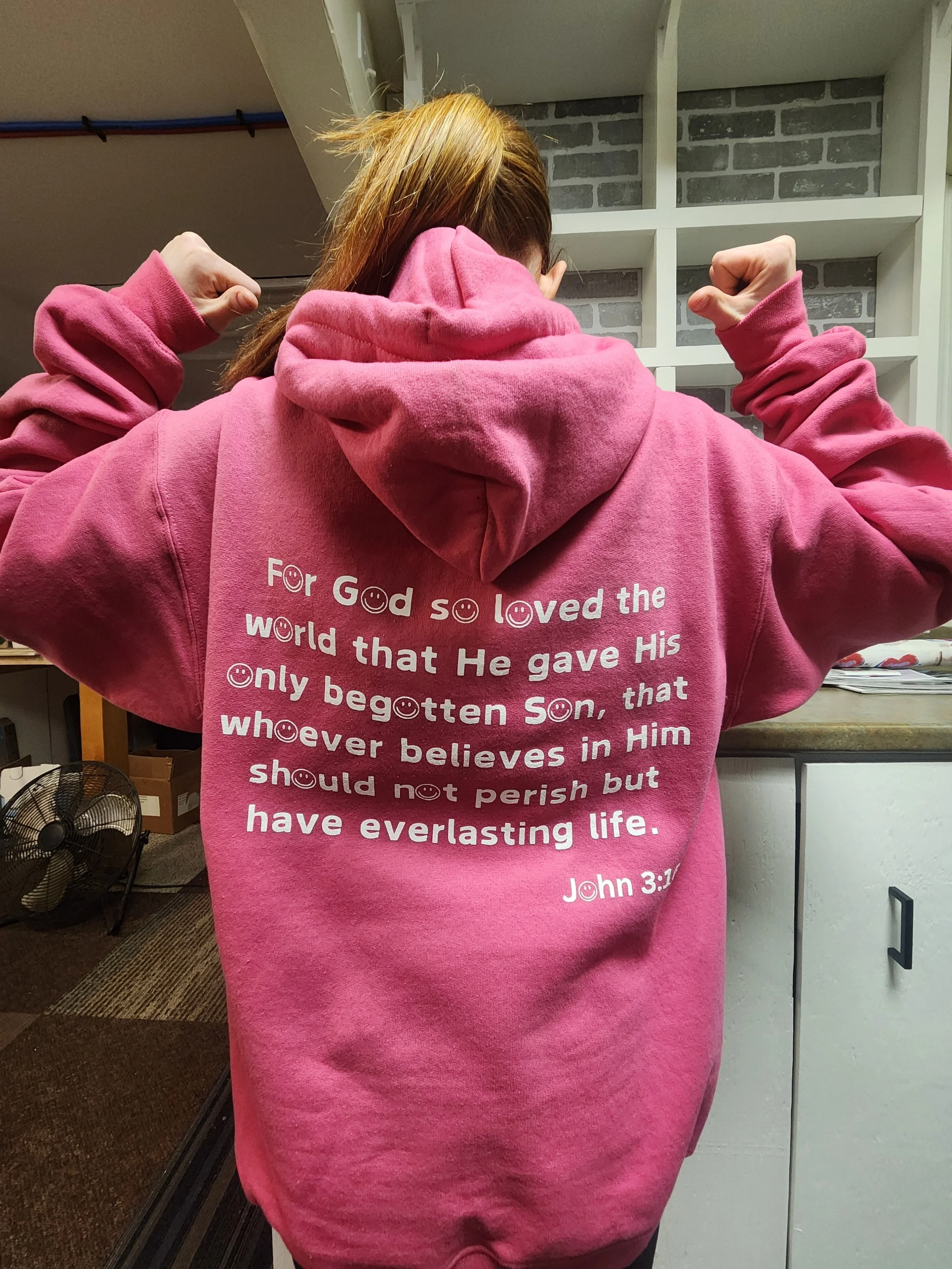 Person in a pink hoodie with their back to the camera. The hoodie has a biblical quote from John 3:16 printed on the back.