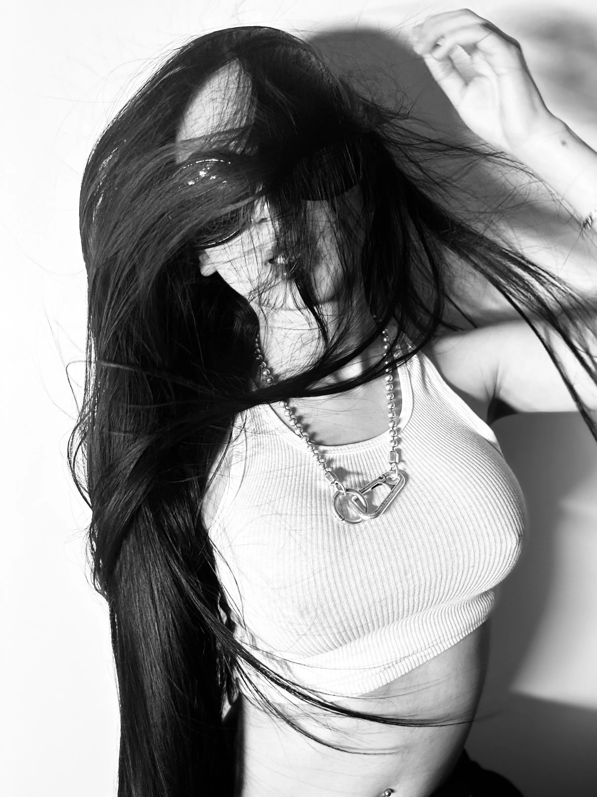 A woman with long dark hair wearing sunglasses, a crop top, necklace, and belly button ring, lying on a plain background with her hair spread out. Styled by PABLO THE STYLIST