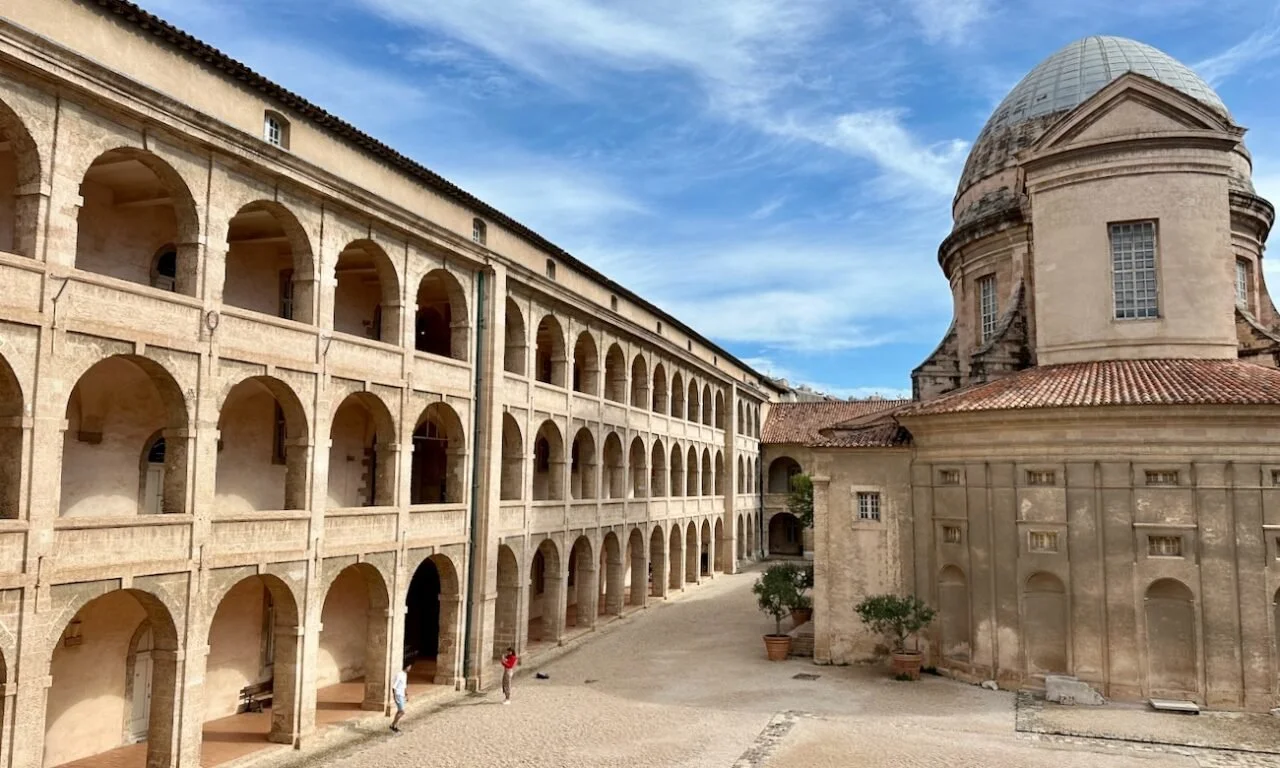 A historic European architectural scene featuring a large building with an arched colonnade and a rounded tower with a domed roof, set against a blue sky with wispy clouds.