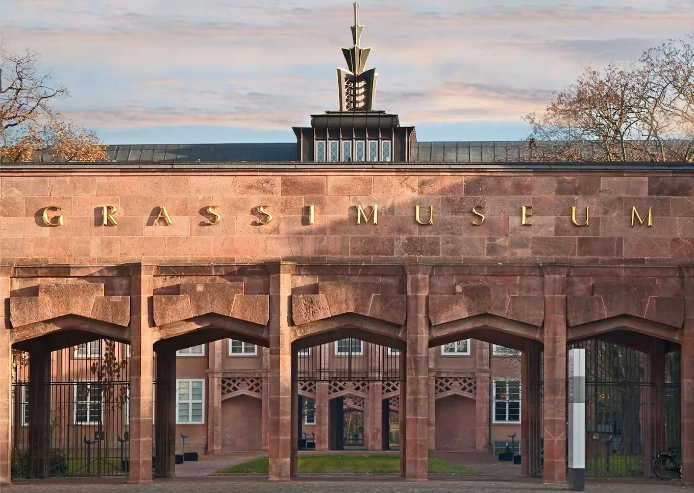 Entrance of the Grassi Museum with a sandstone facade and gold letter signage, trees and a blue sky in the background.