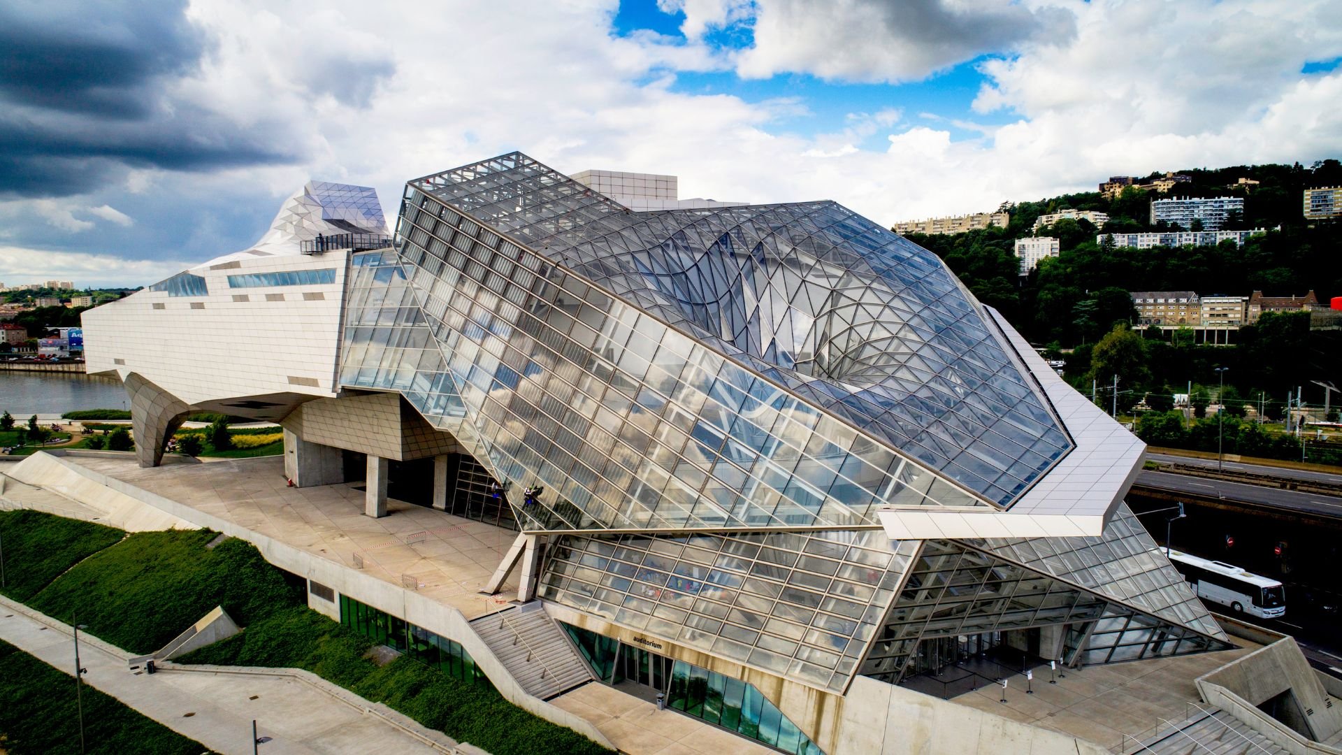 Modern architectural building with angular glass panels, set against a partly cloudy sky, with cityscape and greenery in the background.