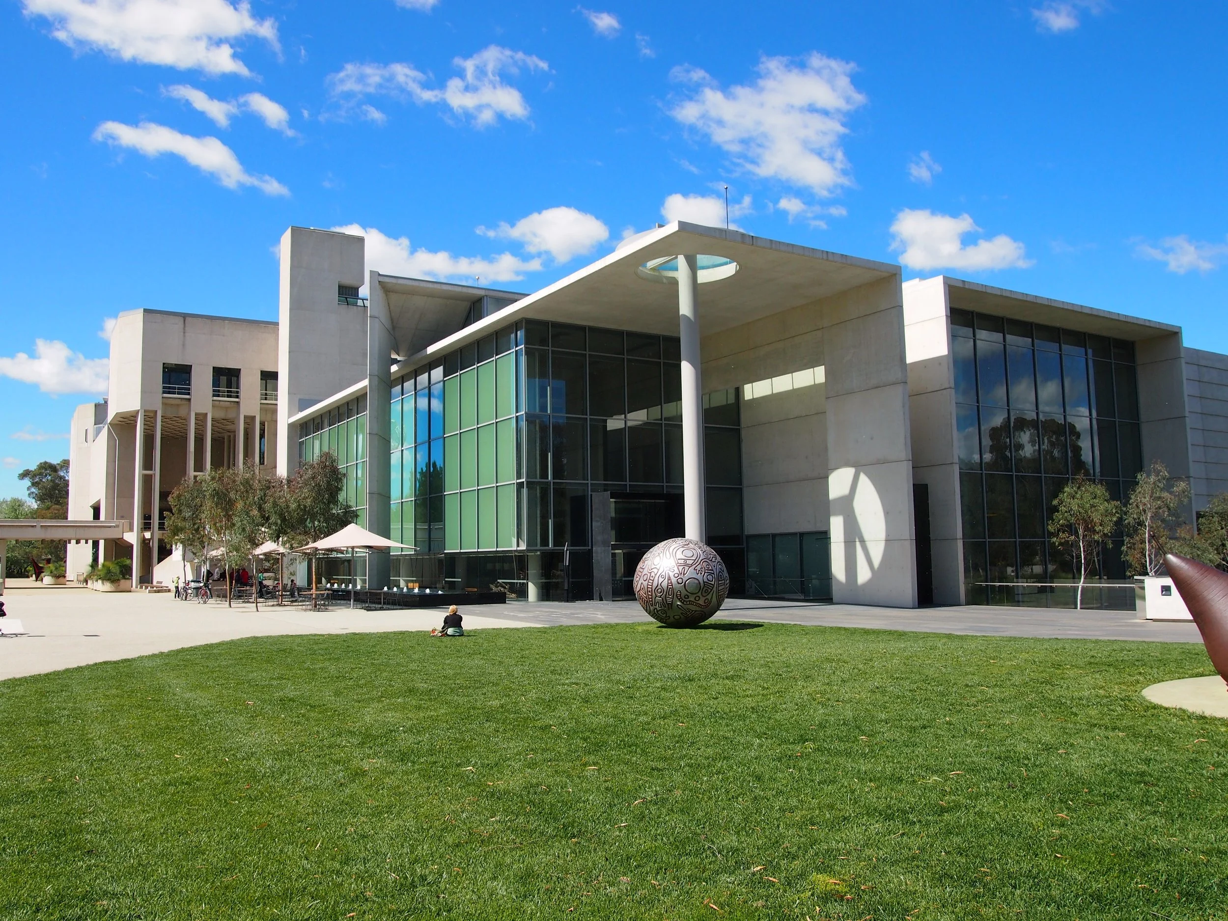 Modern building with large glass windows, a grassy area in front, sculptures, and a few trees against a blue sky with some clouds.