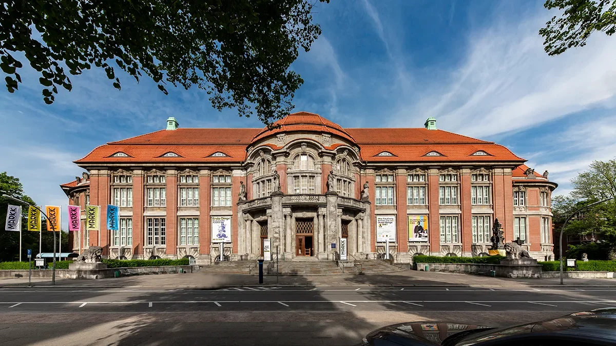 Large historic building with a red roof, detailed stone facade, and flags in front, surrounded by trees and blue sky.