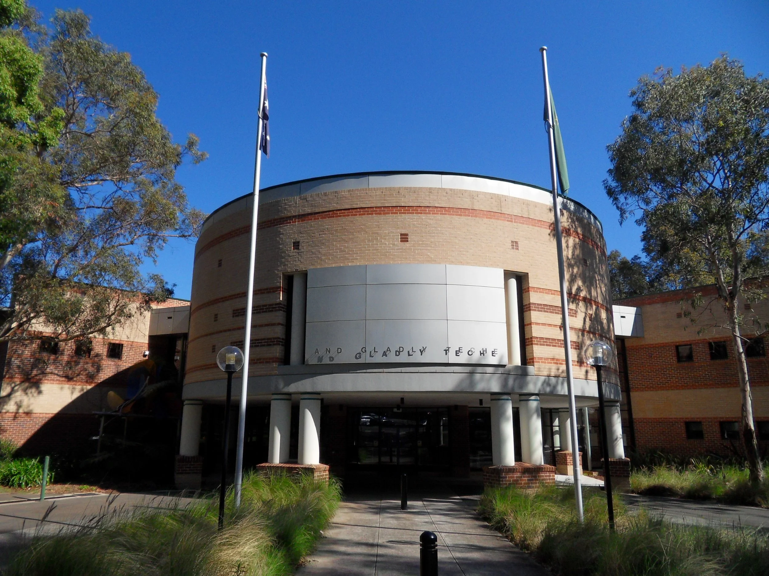 A modern brick building with a rounded facade and tall white columns at the entrance, flanked by two flagpoles and surrounded by trees and bushes, against a clear blue sky.