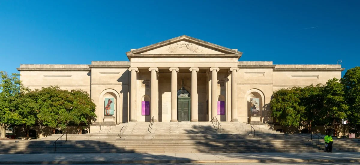 Front view of a neoclassical museum building with large columns, stairs, and banner signs, surrounded by trees, under a clear blue sky.