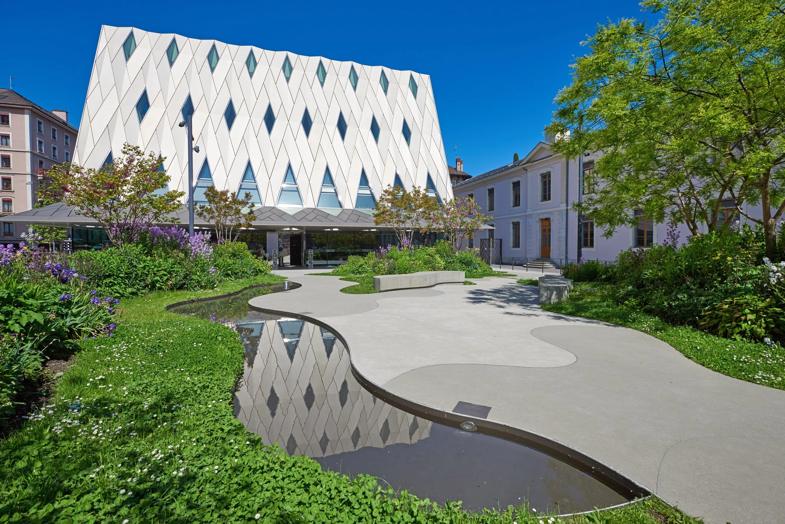 Modern building with a white, geometric, diamond-patterned facade, surrounded by landscaped garden with flowering bushes, trees, and a winding water feature under a clear blue sky.