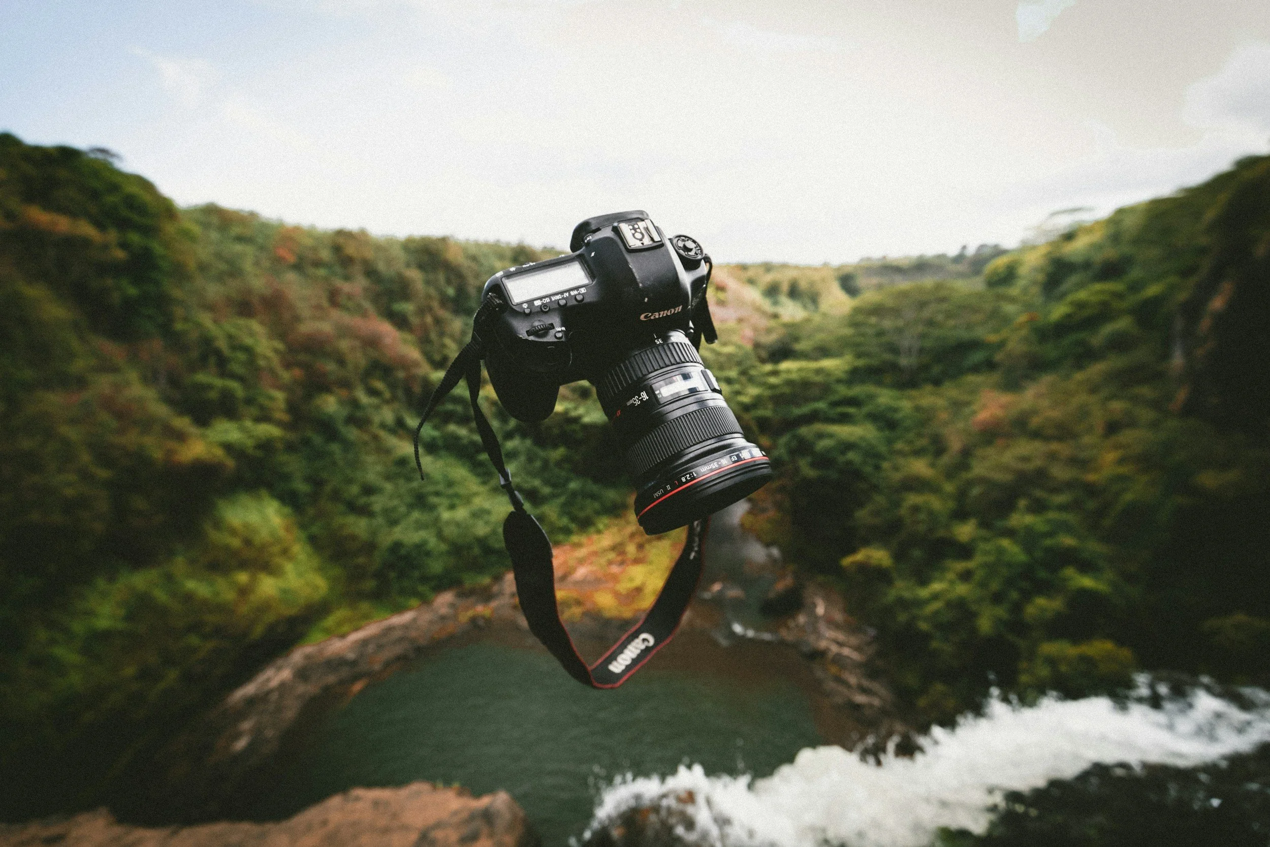 A Canon DSLR camera with a large lens floating in mid-air against a scenic background of a river surrounded by lush green trees and hills.