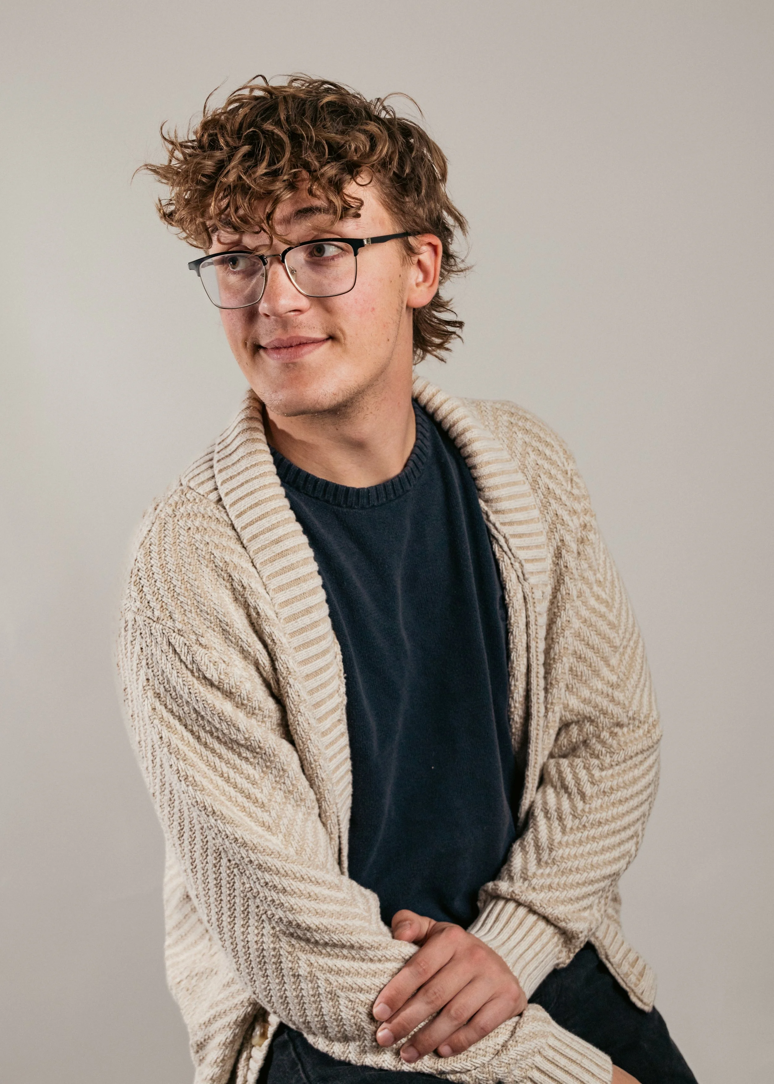 A young man with curly hair and glasses, wearing a beige knitted cardigan over a navy blue shirt, sitting against a plain white background, looking to his left.