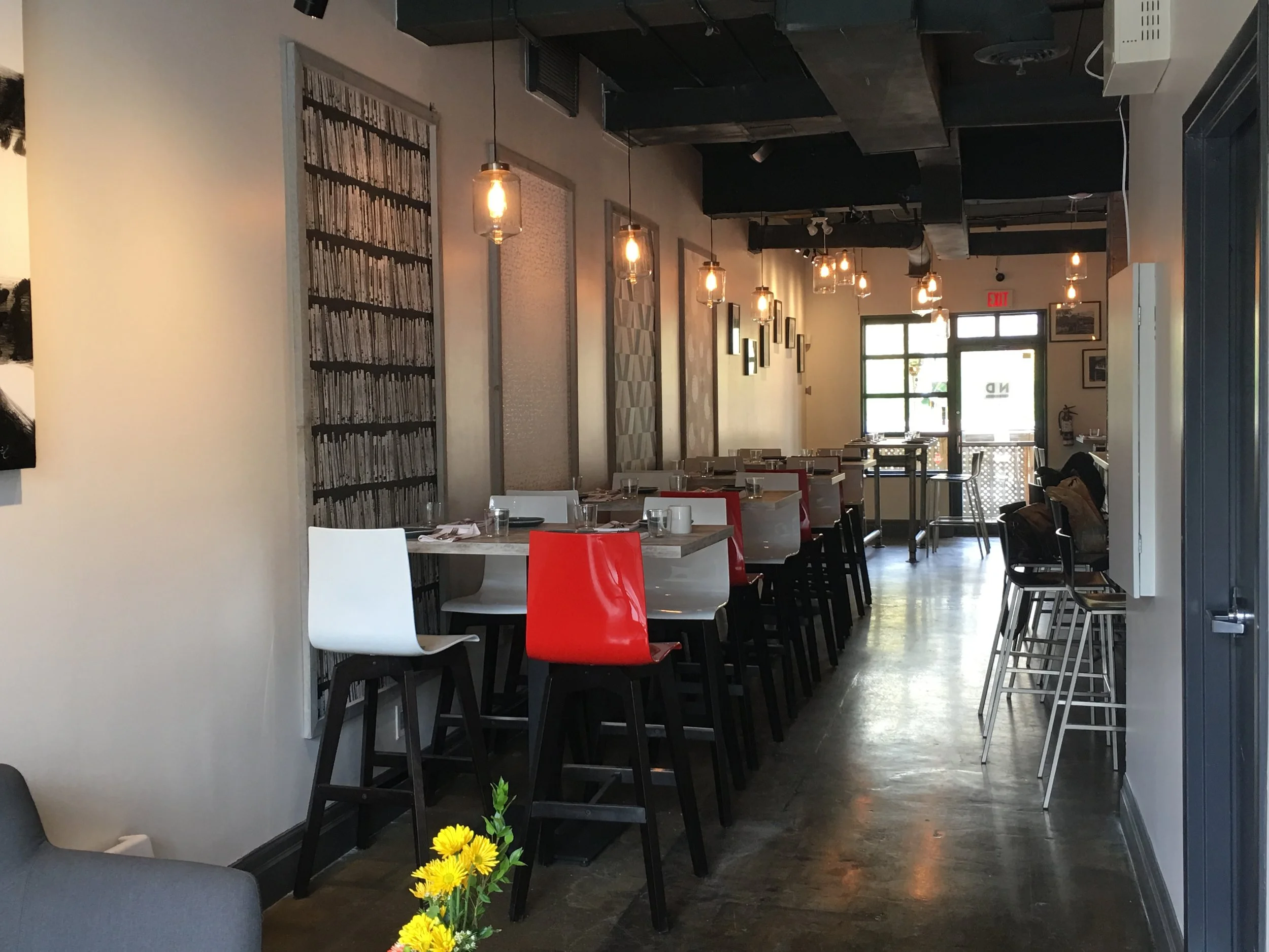 Back dining area at Next Door Restaurant showcasing red and white high chairs, hanging pendant lights, and concrete flooring.