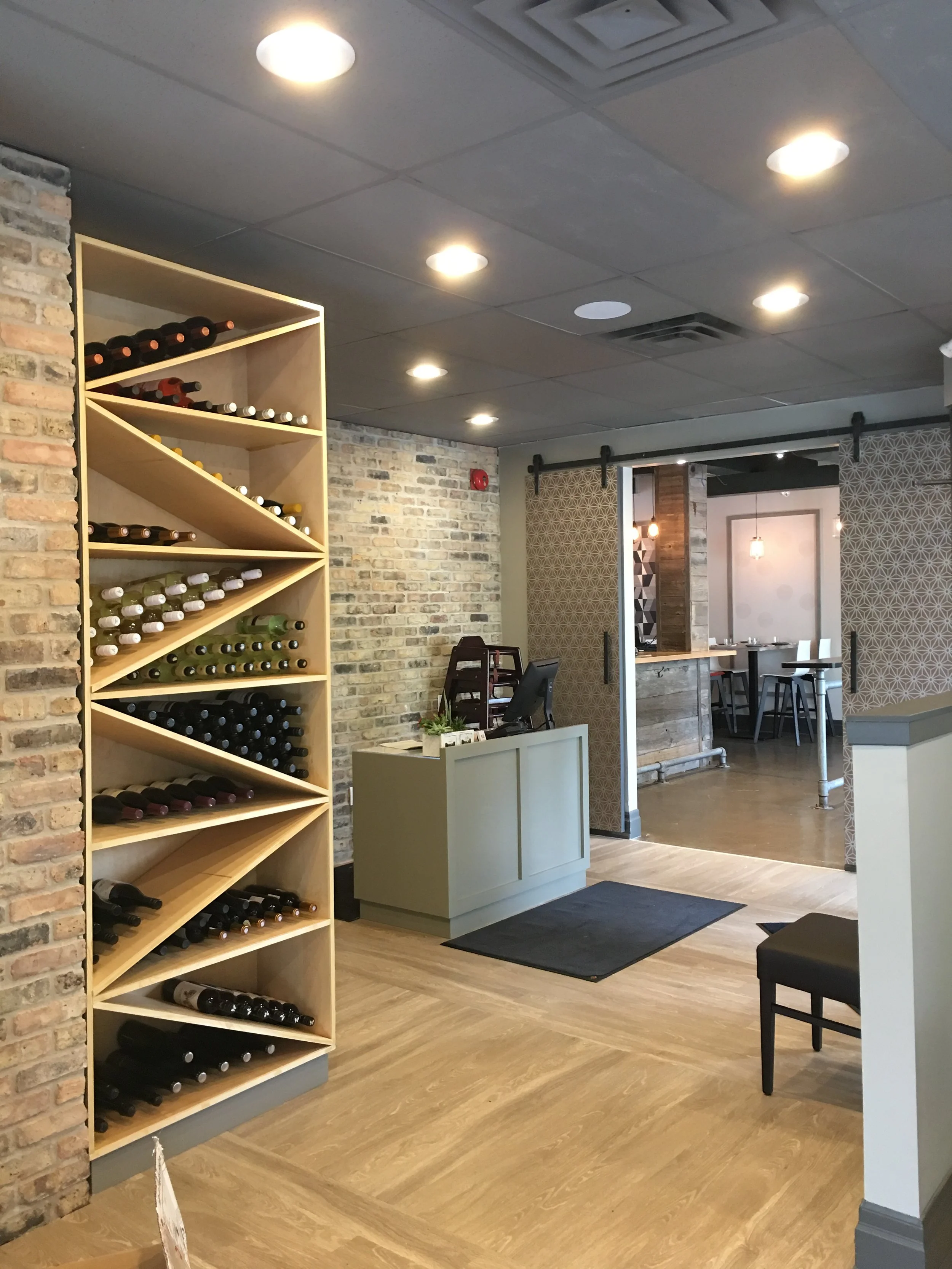 Reception area at Next Door Restaurant featuring diagonal wine storage shelves, brick walls, and wood-patterned flooring.