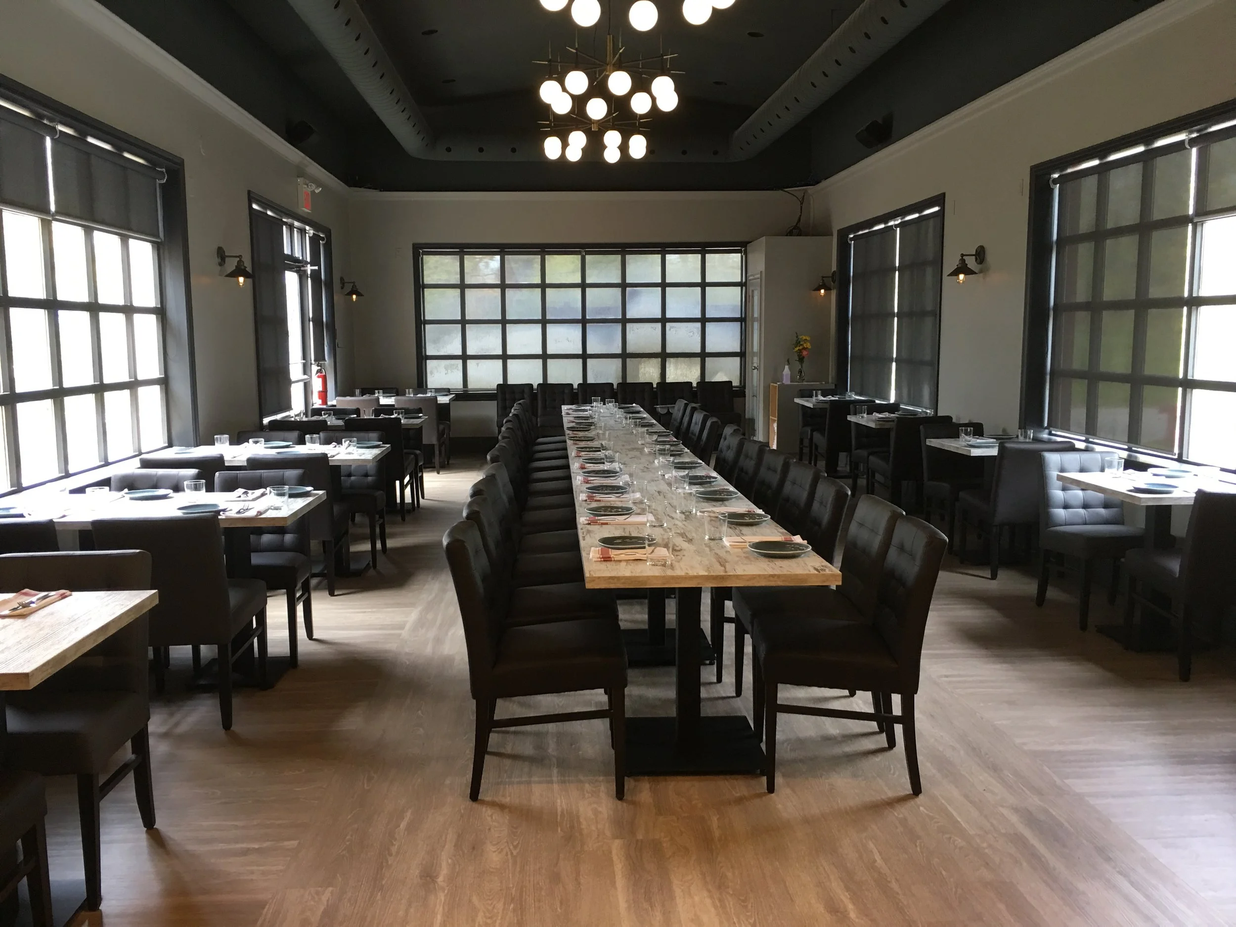Elegant dining room setup with a long communal table, modern chandeliers, and grid window design at Next Door Restaurant.