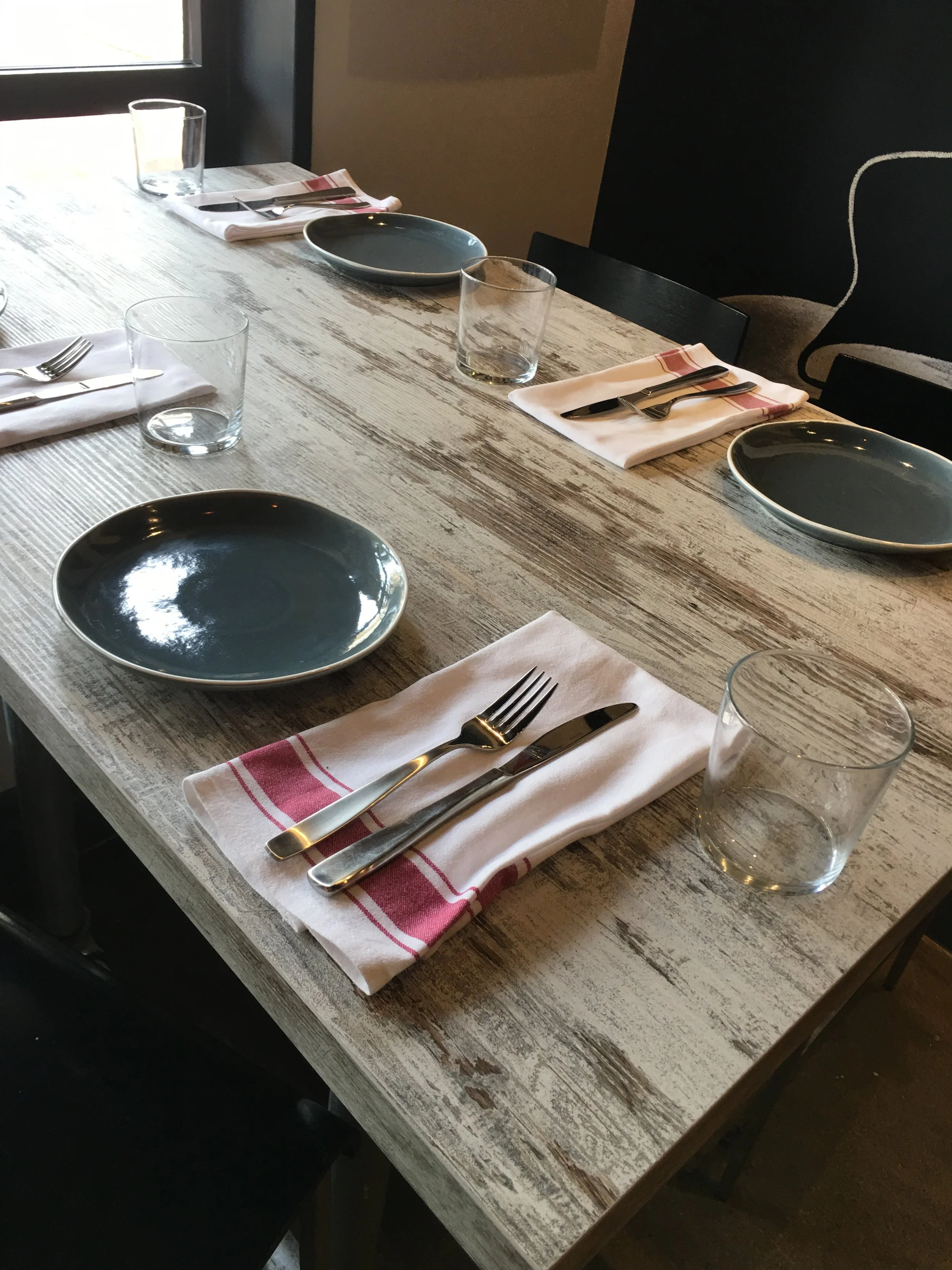 Rustic wooden table at Next Door Restaurant set with blue ceramic plates, clear glasses, and neatly folded napkins.
