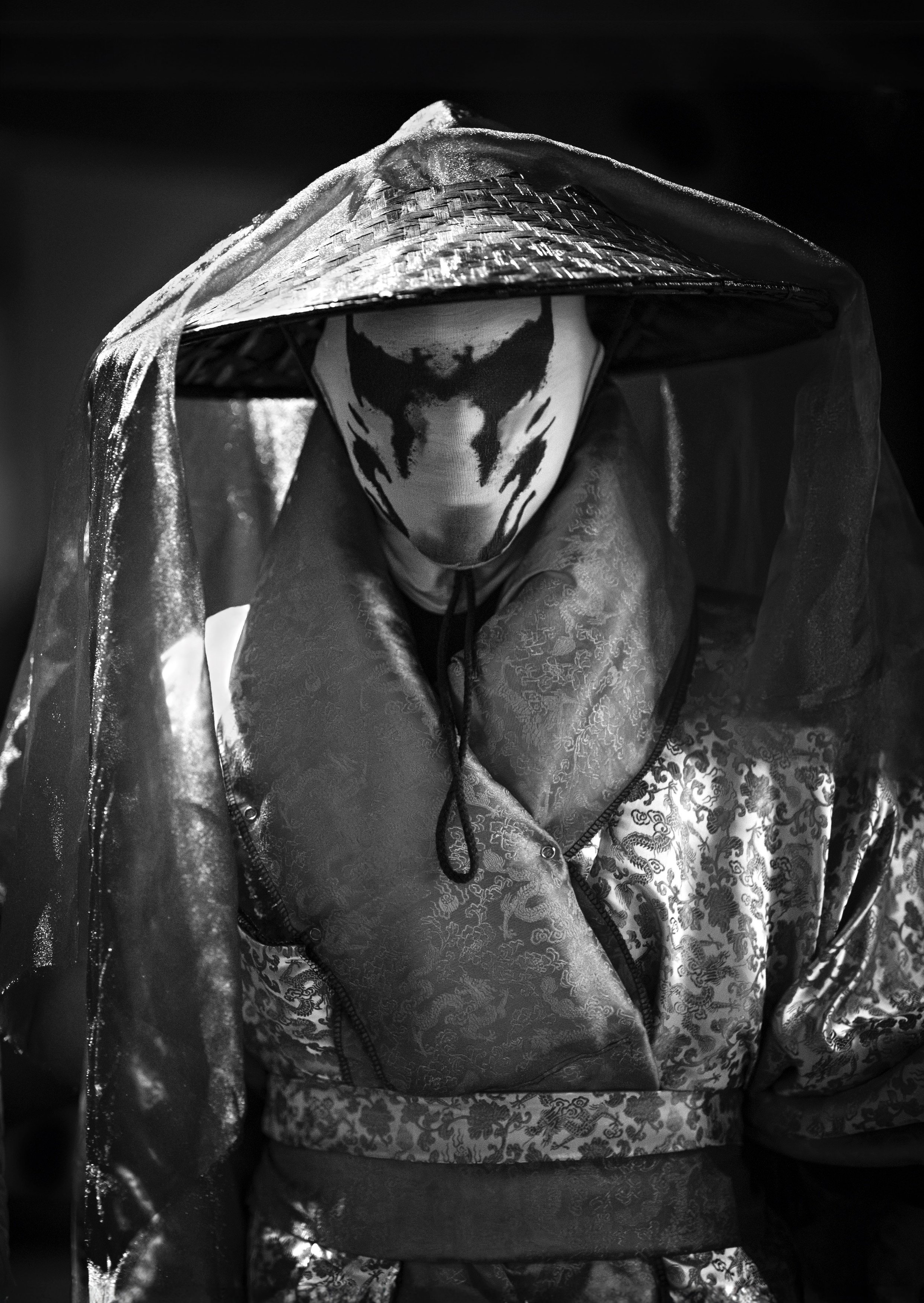 Person wearing a patterned cloak and hat with a veil, face obscured by a mask with inkblot design, black and white image.