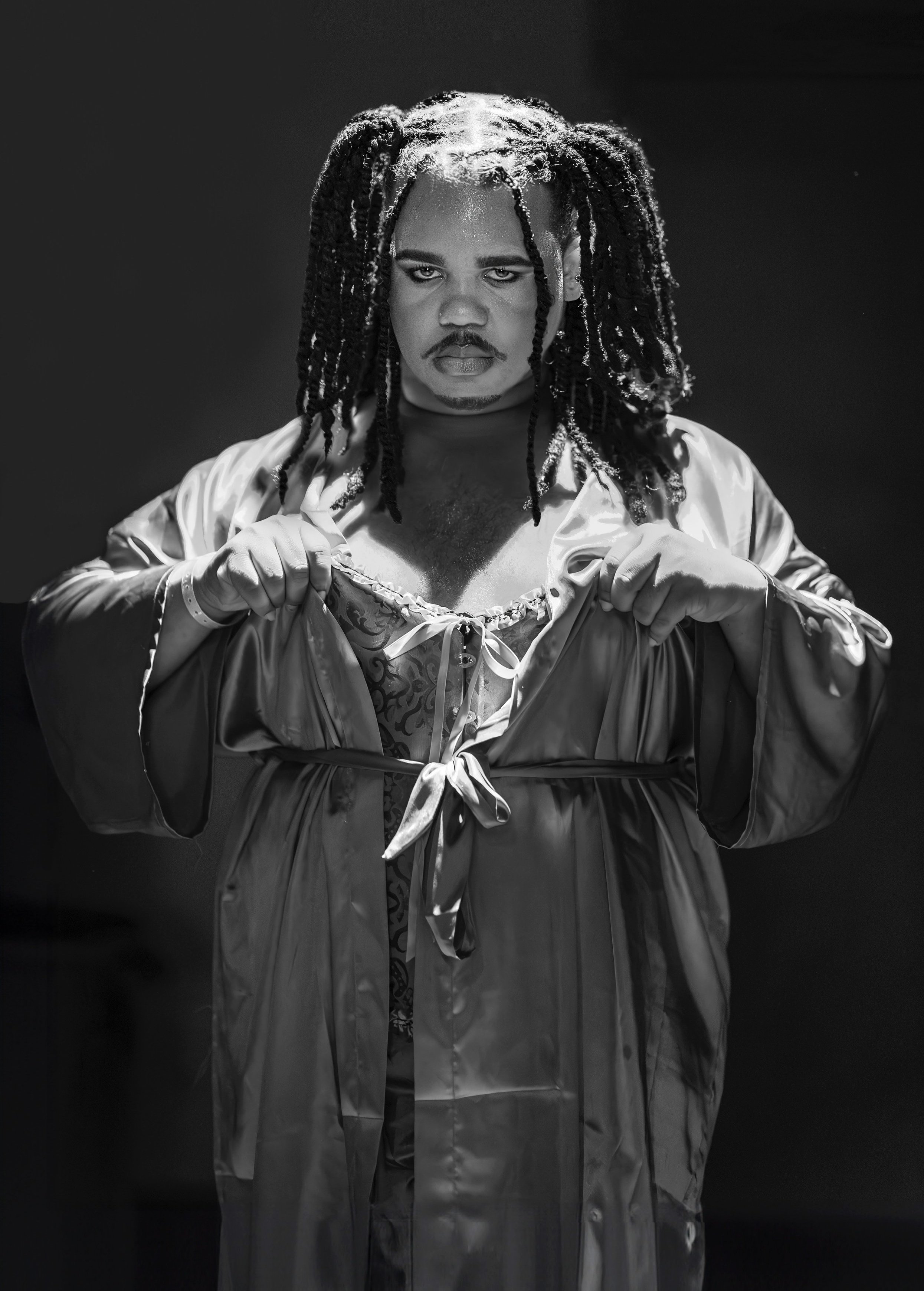 Person with braided hair wearing a robe and lace attire, posing in dramatic lighting, black and white. Image by portrait photographer.
