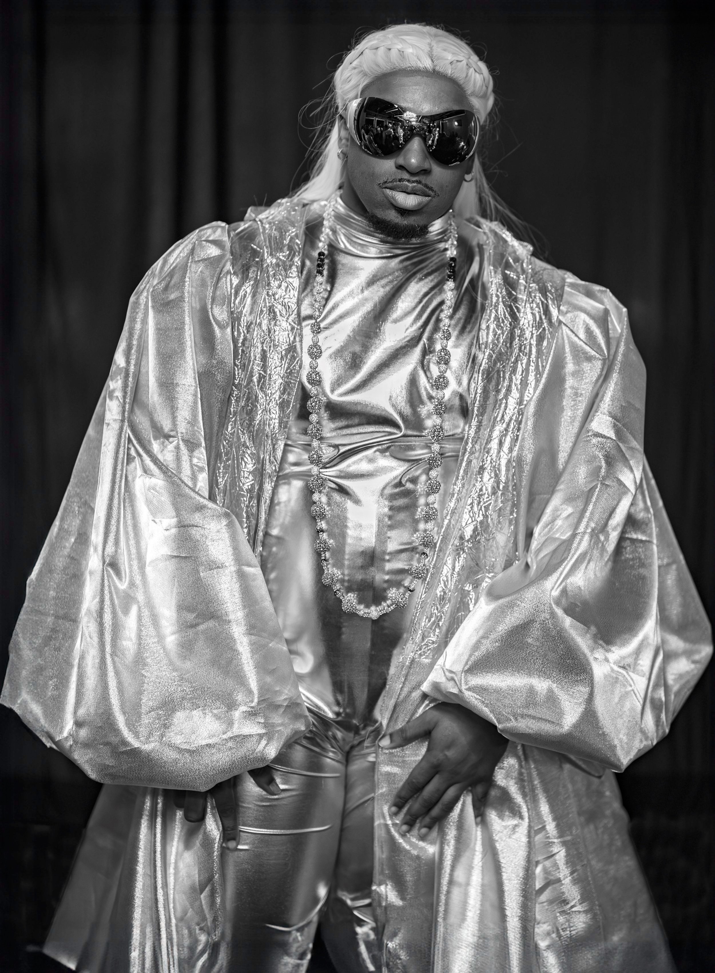 Person in metallic silver outfit with long sleeves and cape, wearing sunglasses and a long necklace.