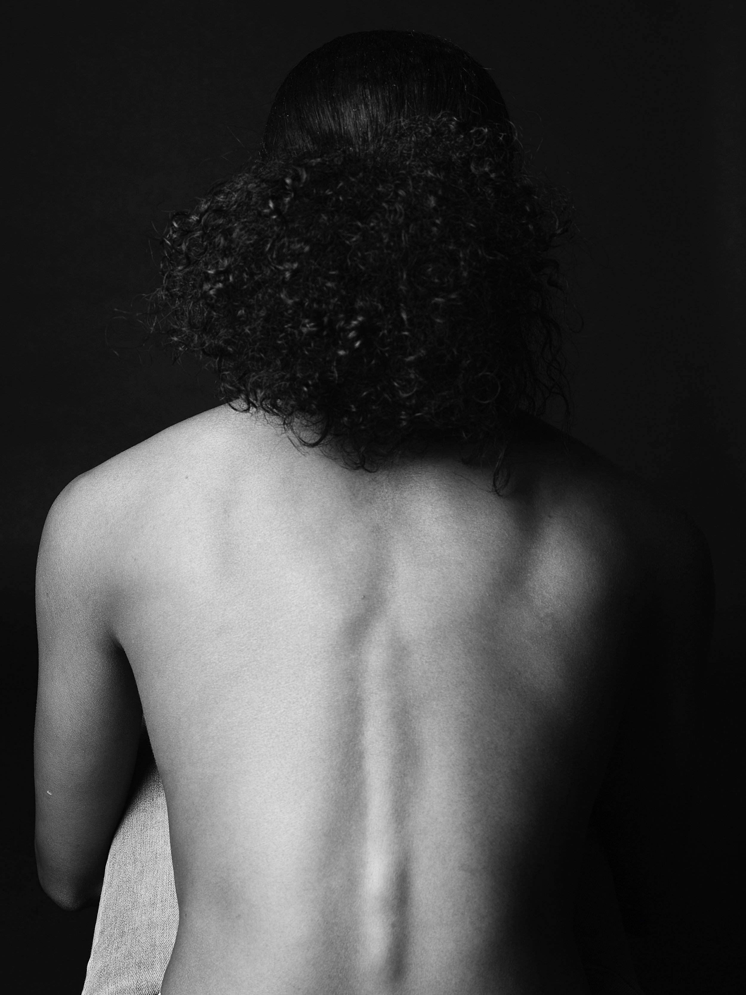 Black and white studio photography image of a person's back with curly hair, sitting against a dark background.