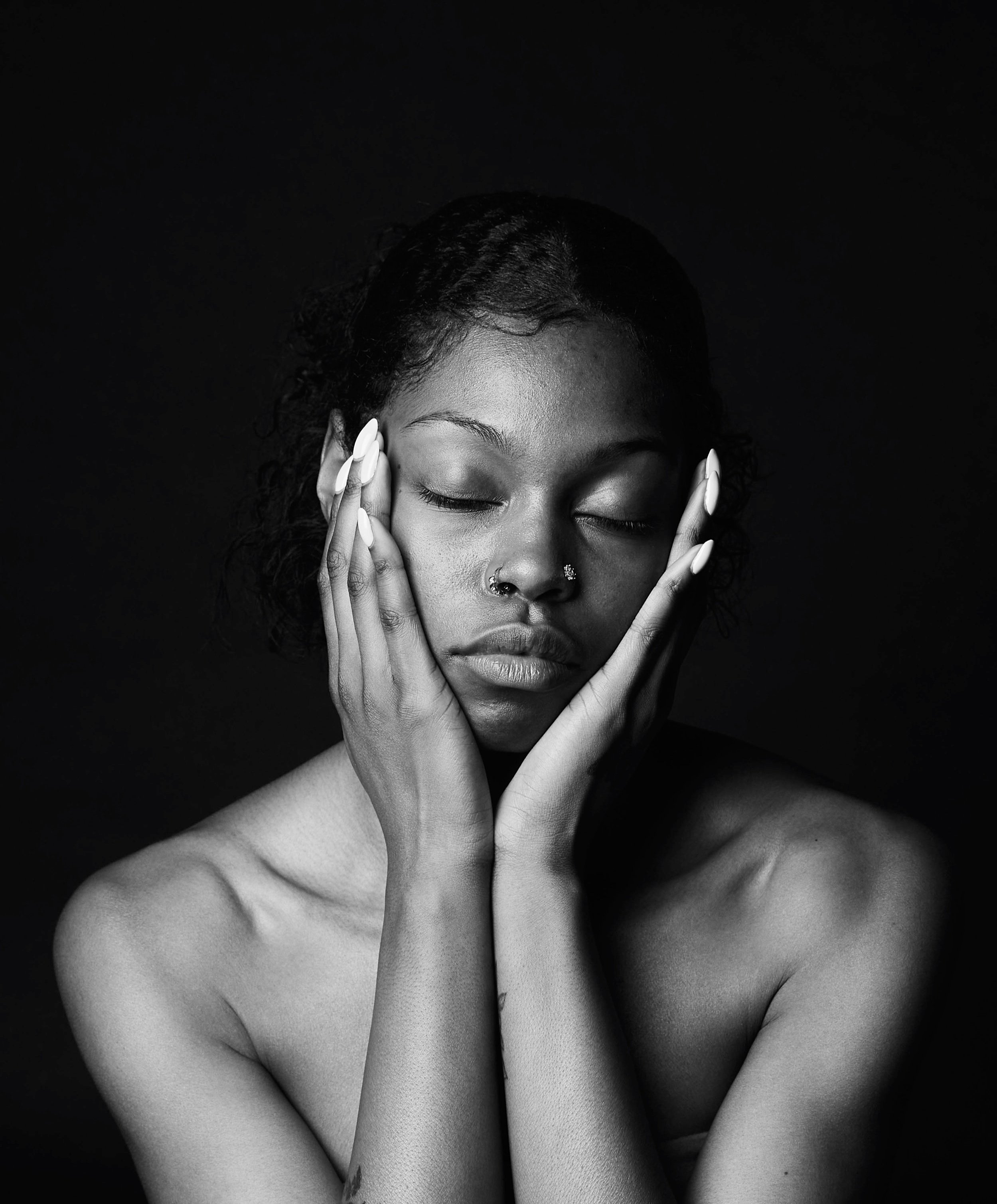 Black and white studio portrait of a woman with closed eyes, hands on face, and nose rings.