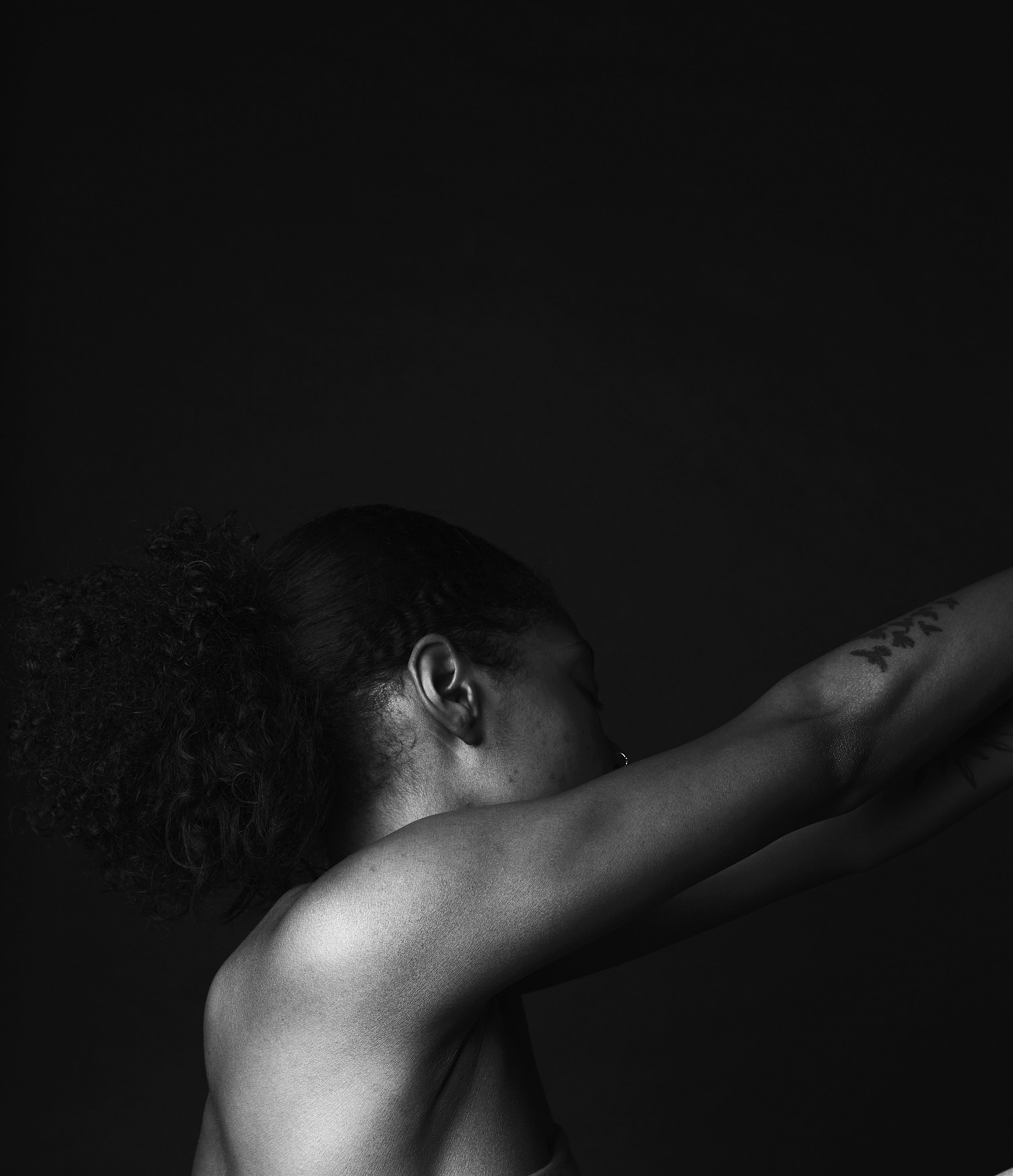 Black and white image of a person with curly hair in a bun, facing away, arm raised, tattoos visible on arm, against a dark background.