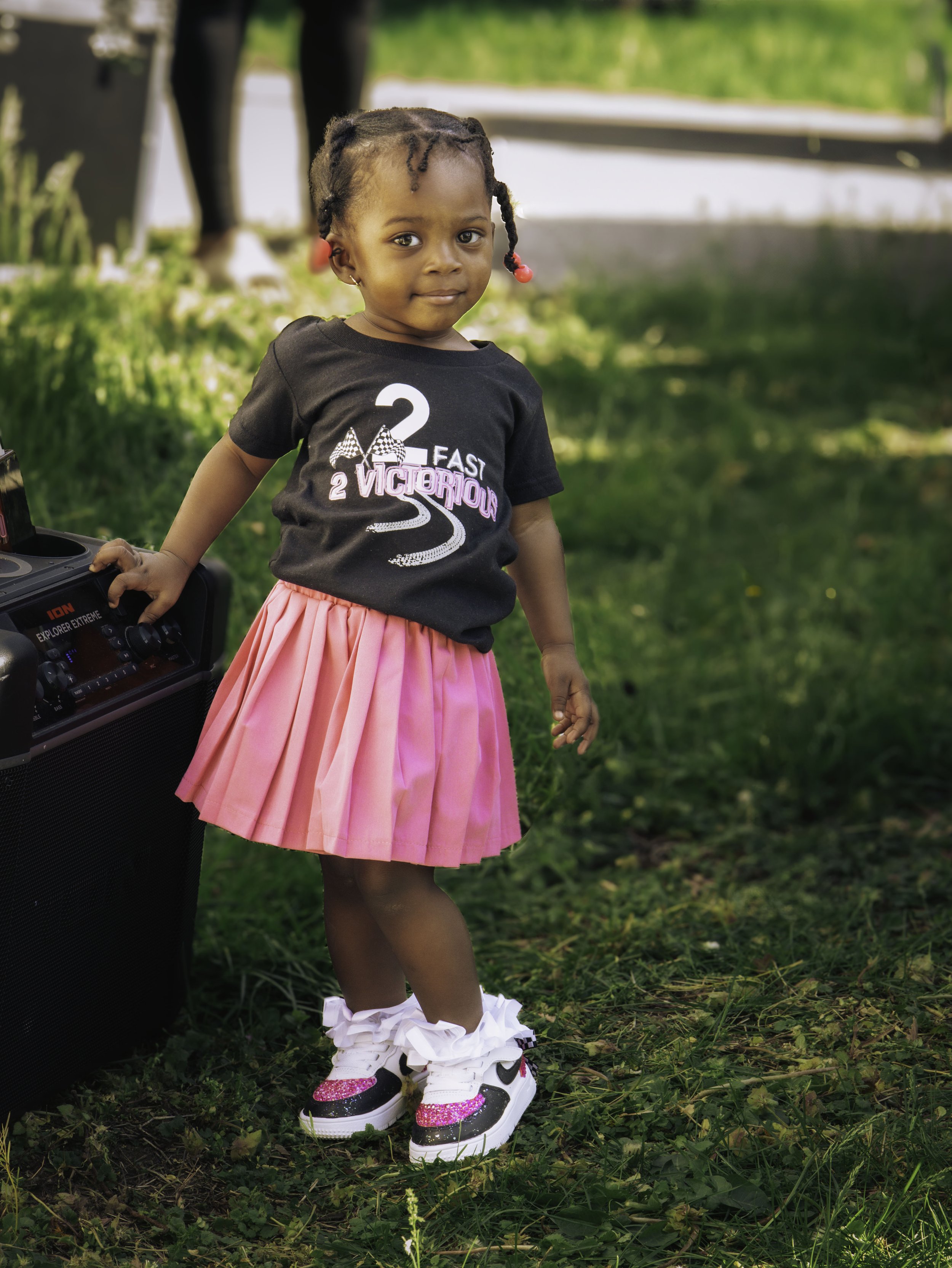Young child with braids wearing a black t-shirt, pink skirt, and stylish sneakers standing on grass next to a speaker. Image by DMV area family photographer.