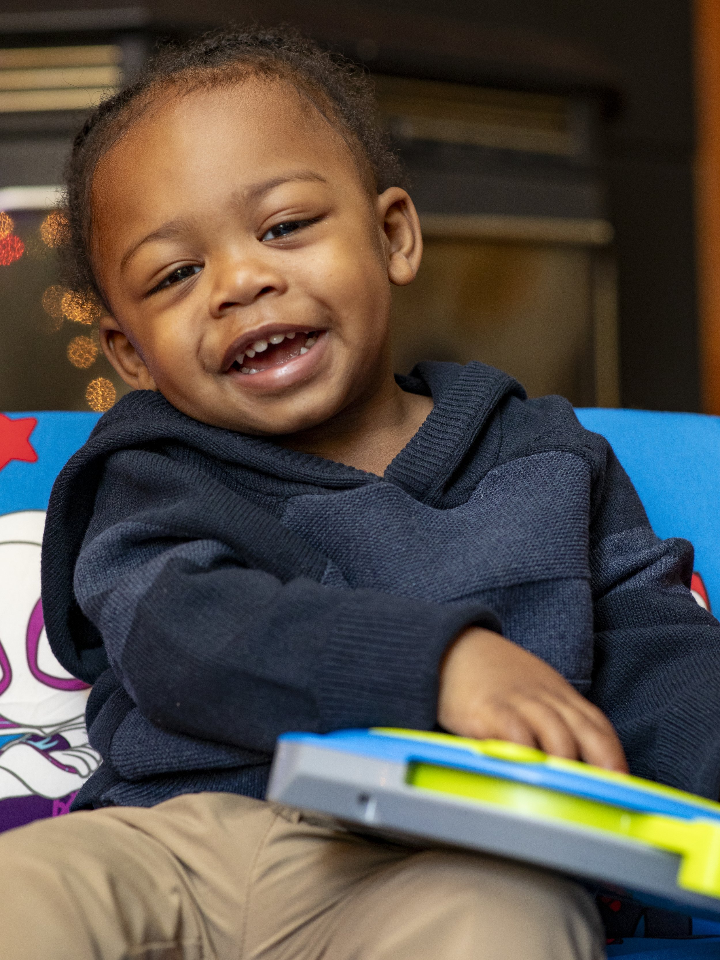 Family photographer captures smiling toddler sitting on a colorful chair, wearing a navy sweater and holding a toy.