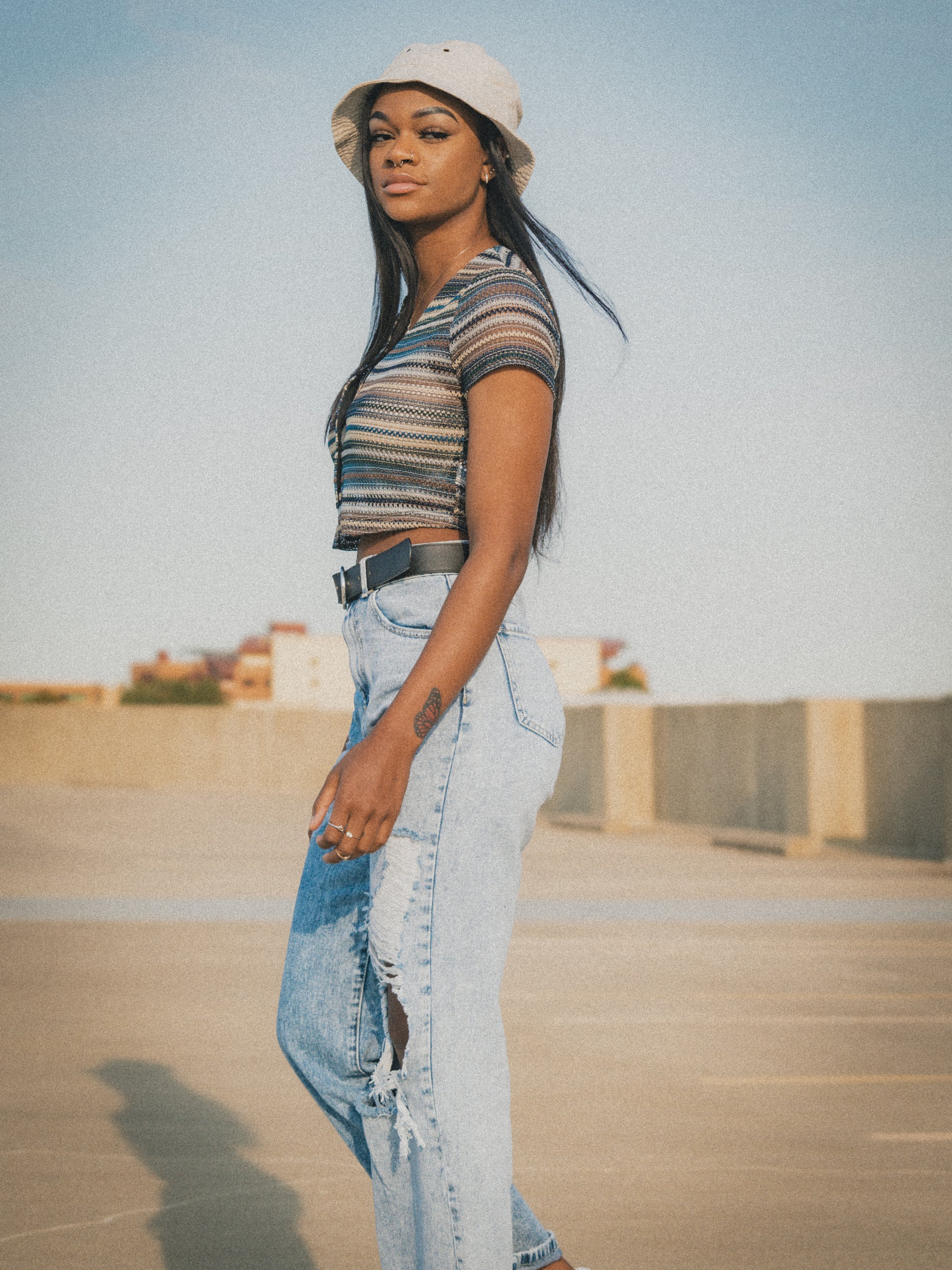 Person in striped shirt, ripped jeans, and beige bucket hat standing on outdoor pavement. Image by Maryland portrait photographer.