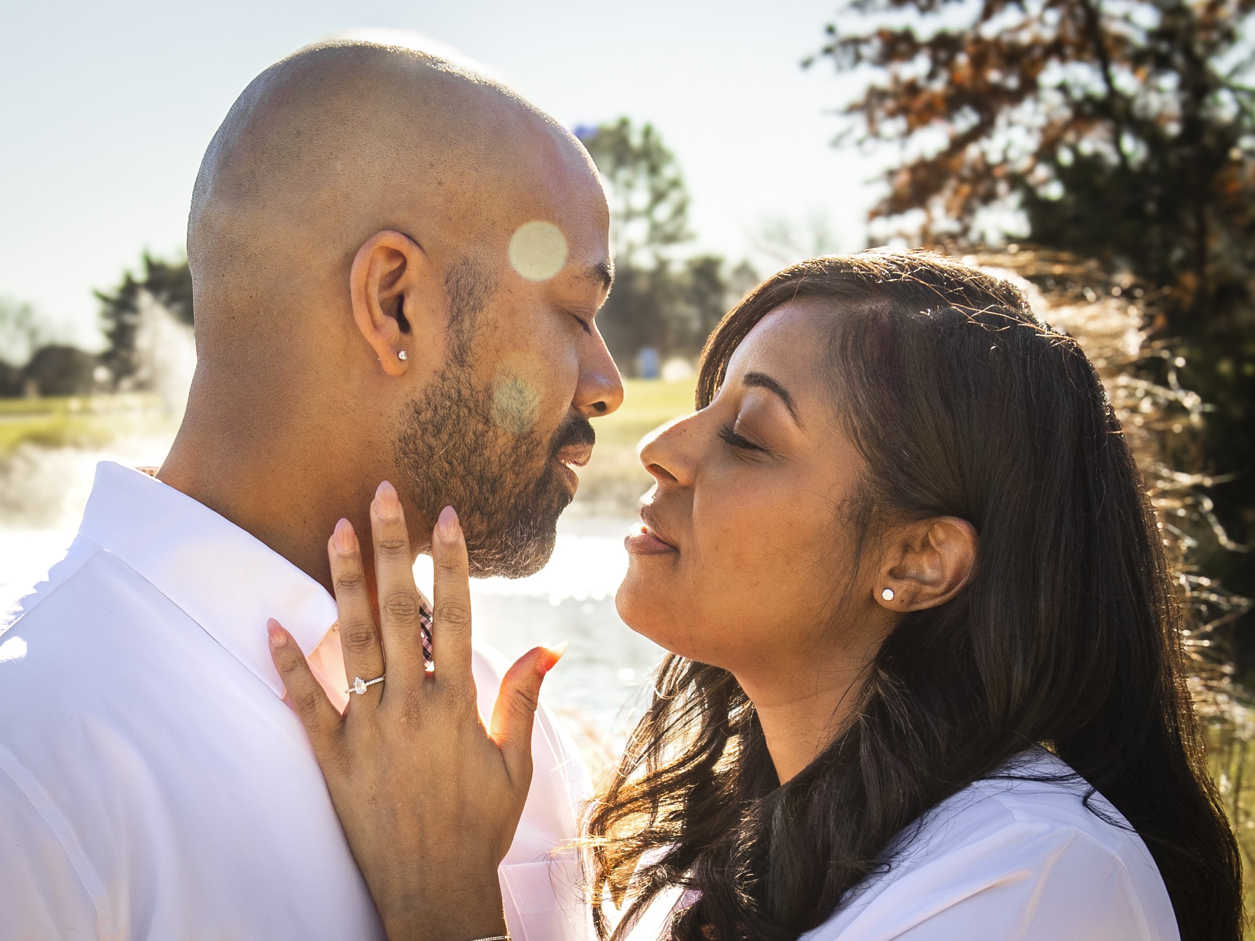 A couple in love face each other with their eyes closed, wearing white shirts, outdoors. The woman displays an engagement ring on her hand as she gently touches the man's face. Image by Maryland portrait photographer.