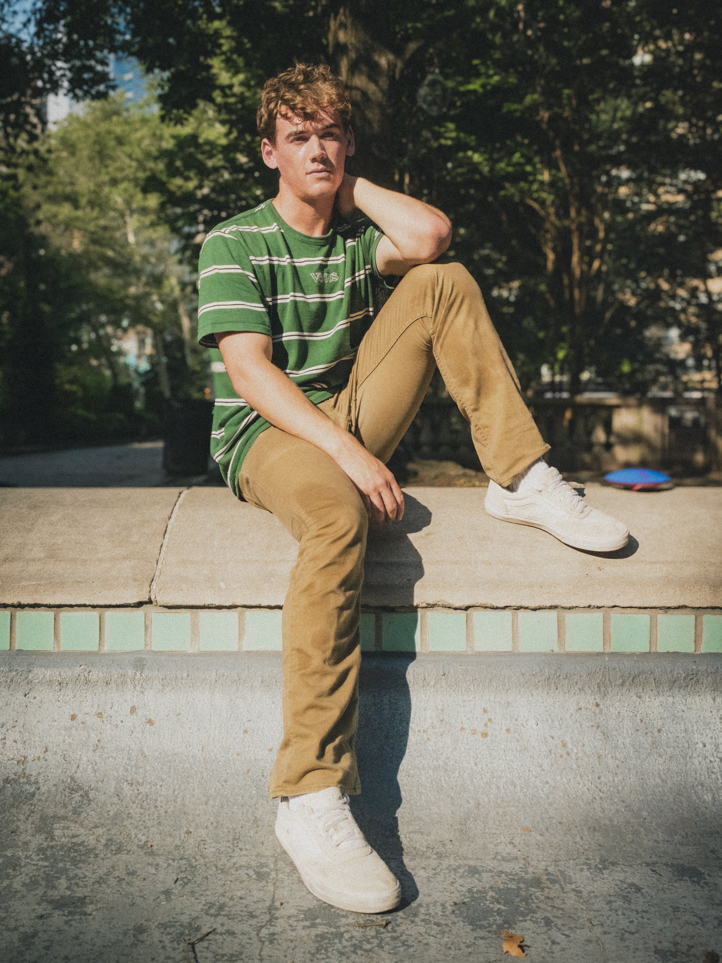 Young man in a green striped shirt and khaki pants sitting on a stone ledge outdoors in a park, with trees in the background. Image by lifestyle photographer.