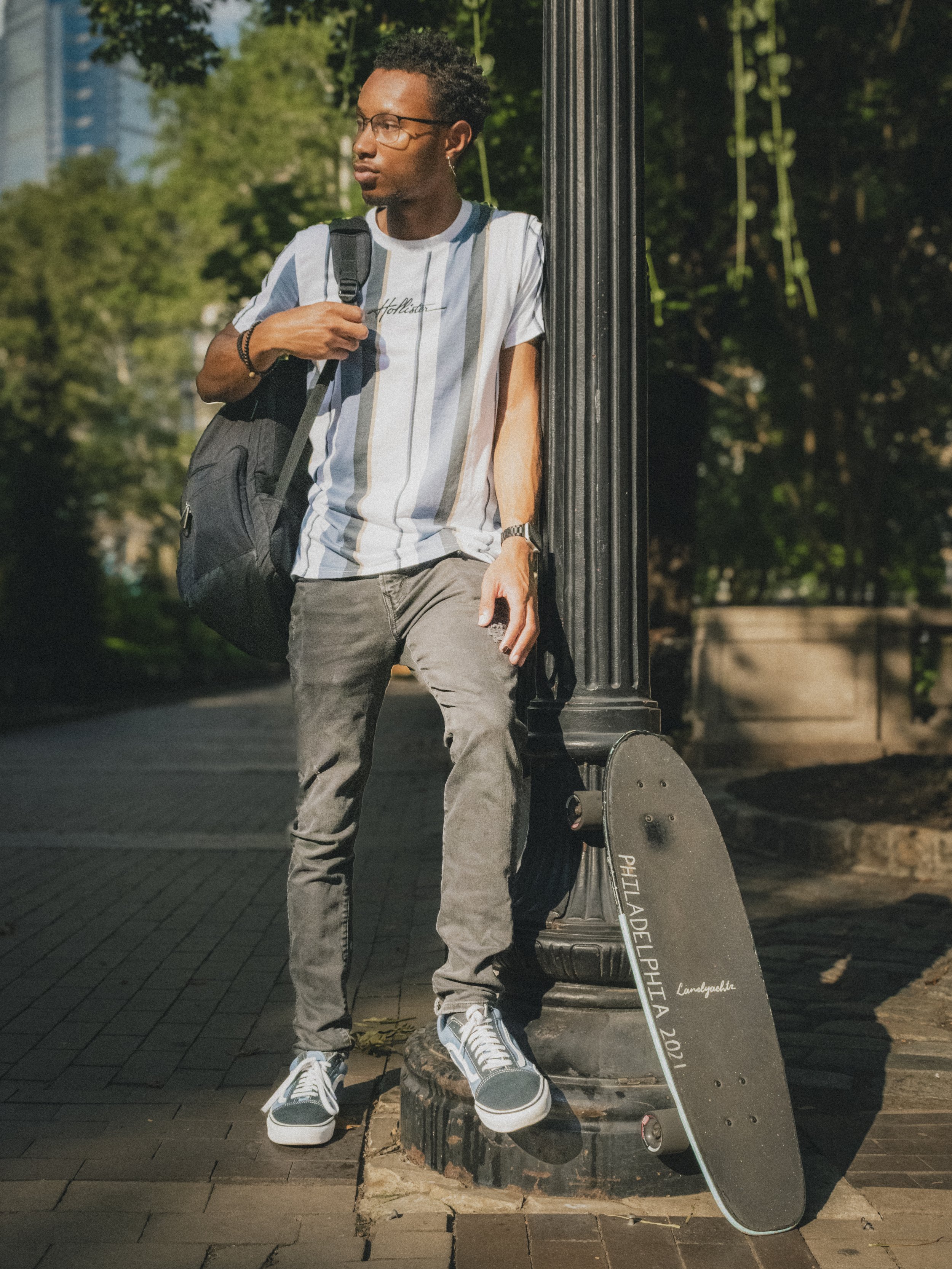 Person leaning against a lamp post with a skateboard nearby, wearing a striped shirt, jeans, sneakers, and a backpack, in an outdoor setting with trees in the background. Image by Maryland lifestyle photographer.