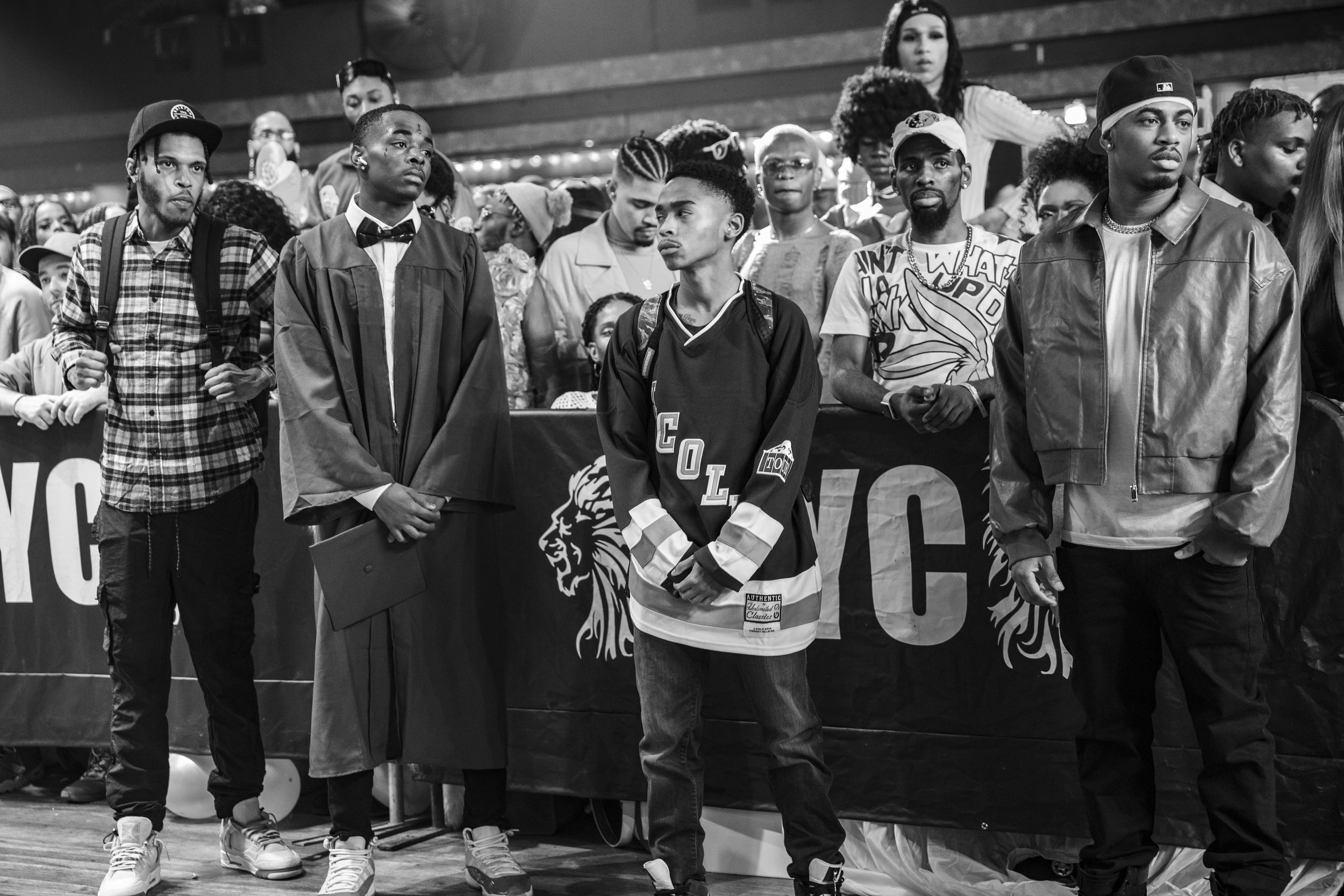 Event photographer captures black and white photo of people standing in a crowd, with one person in a graduation gown and others in casual attire, including a hockey jersey and a leather jacket.