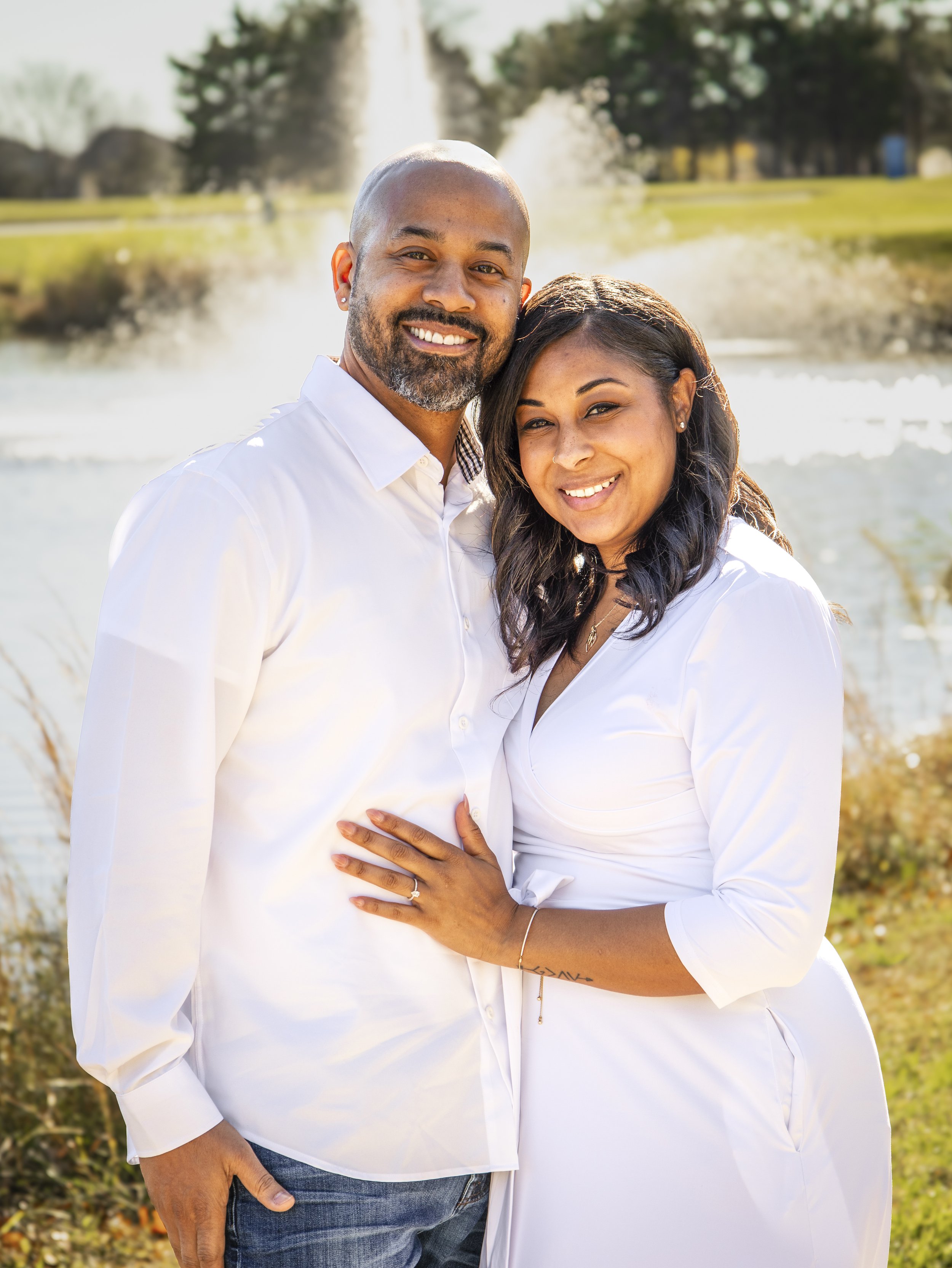 A couple wearing white clothing, embracing and smiling, standing outdoors in front of a pond with a fountain. Image by Maryland family photographer.