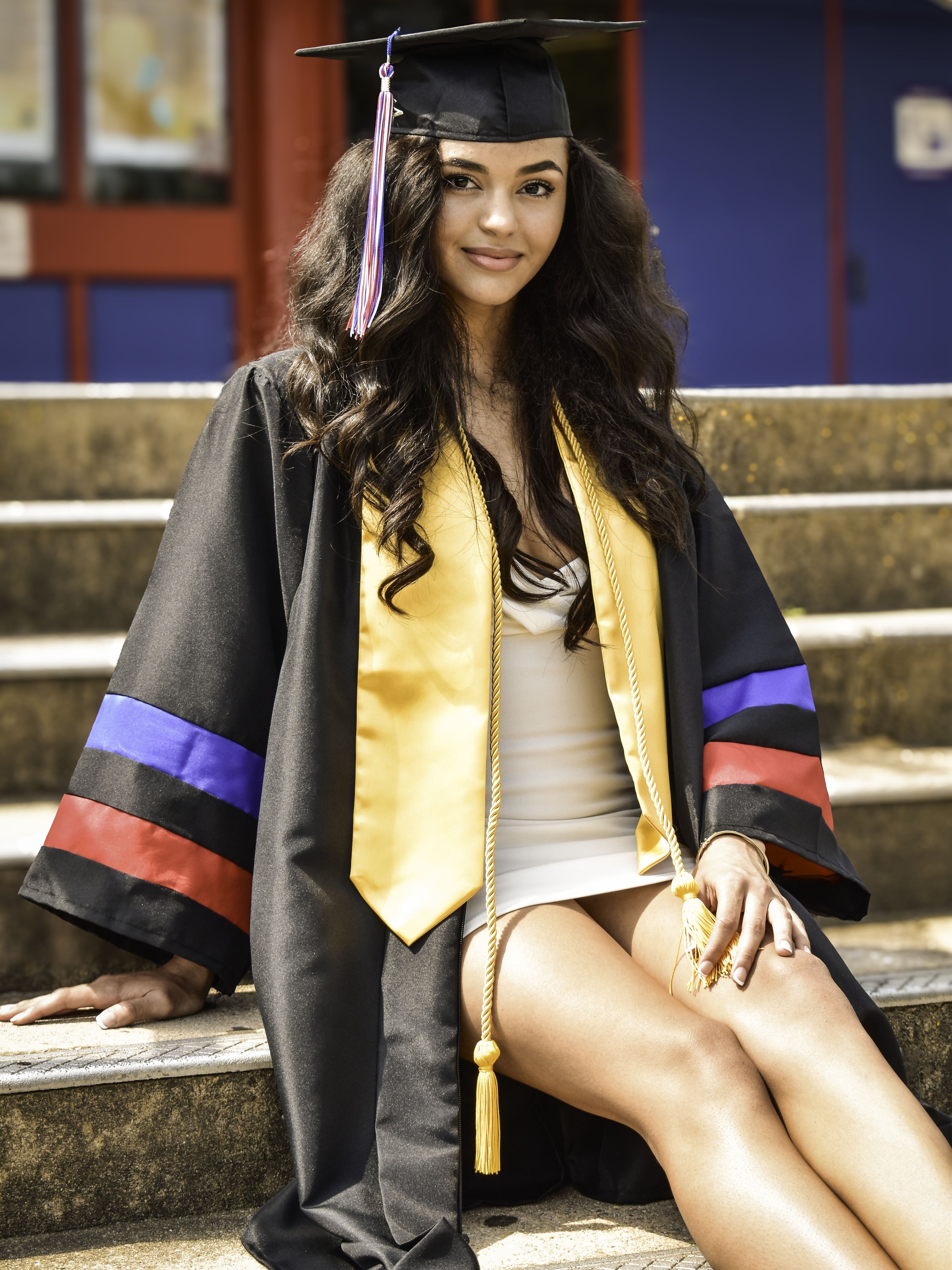 A young woman in a graduation cap and gown sits on outdoor steps. She wears a black robe with purple and red stripes on the sleeves and a yellow stole. Her long hair is loose and she smiles at the camera.