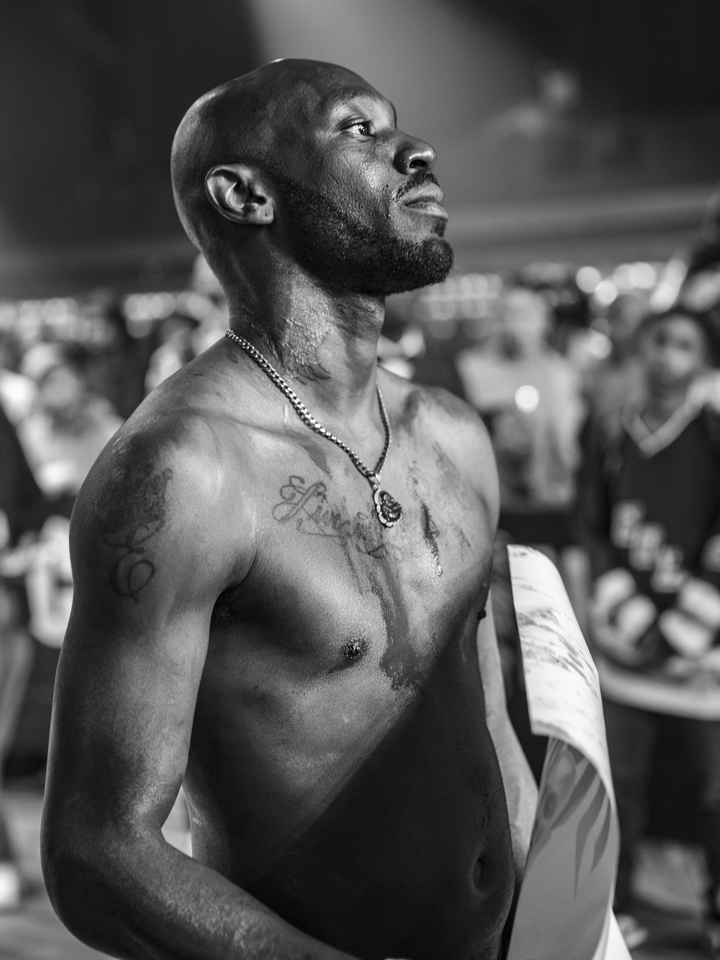 Black and white photo of a shirtless man with tattoos and a necklace, standing in an event crowd, looking into the distance. Image by event photographer.