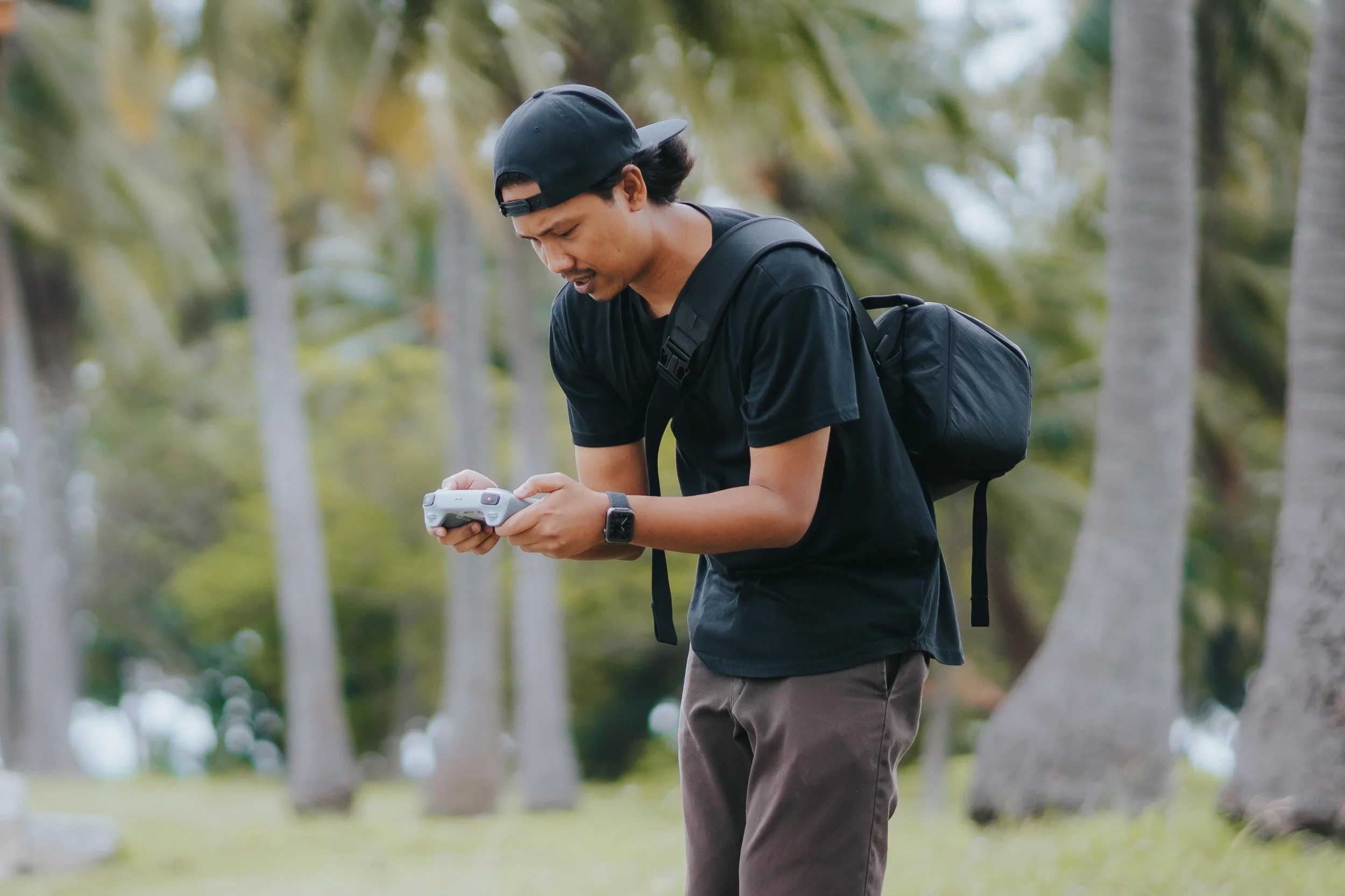 A man wearing a black cap, black t-shirt, and dark pants is standing outdoors during daytime, looking down at a drone in his hands. He has a black backpack and a watch on his left wrist, with a background of green trees.