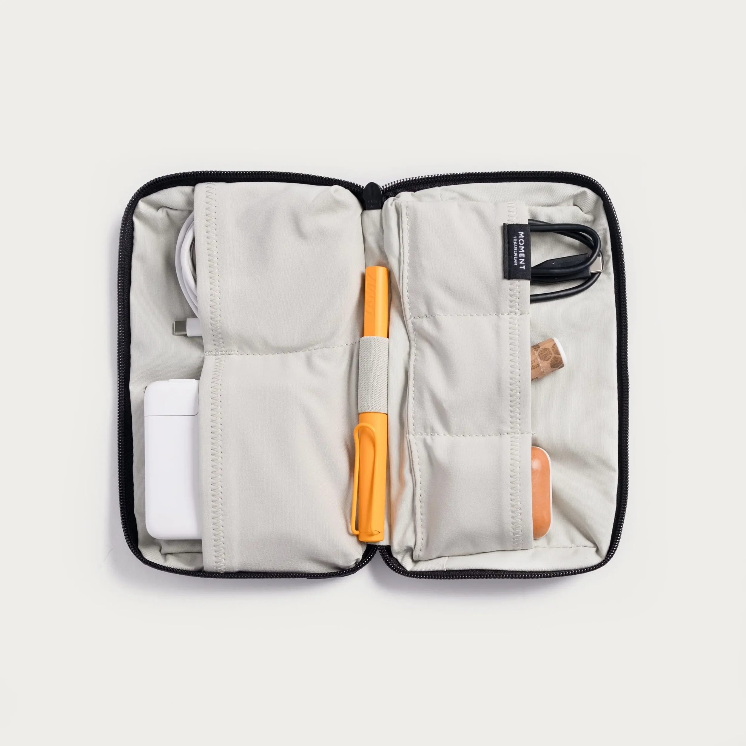 A travel kit organizer on a white background with compartments holding various travel essentials including a white charging cable, a black charging cable, an orange highlighter, and a small cork object, all inside a beige fabric case with black zipper.