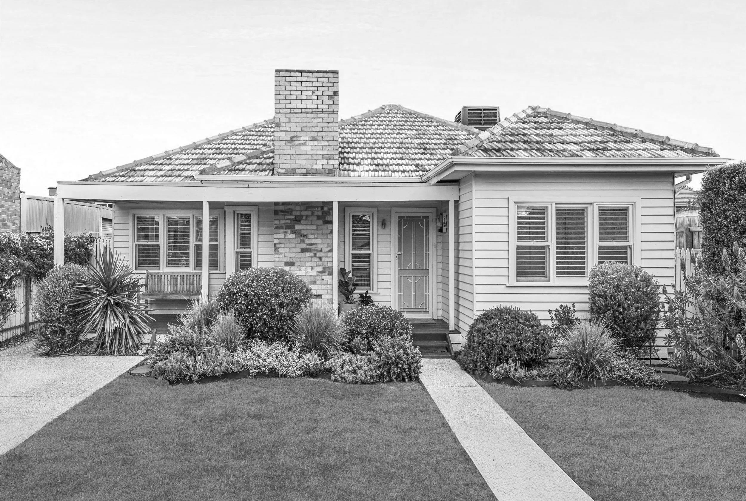 Black and white photo of a suburban house with a brick fence, trees, bushes, a front yard, and a sidewalk.