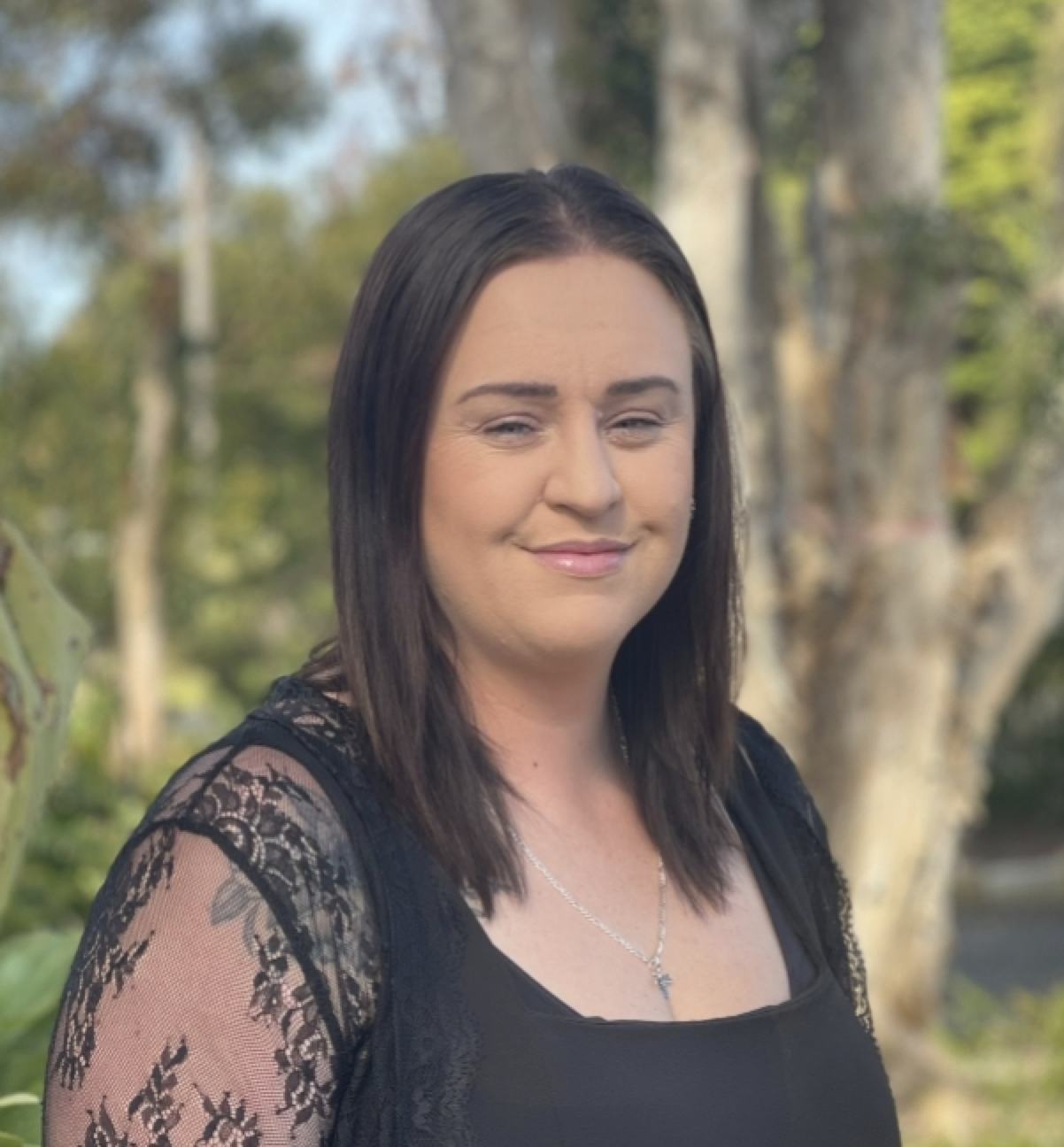 A woman with shoulder-length dark brown hair and light skin is smiling outdoors.