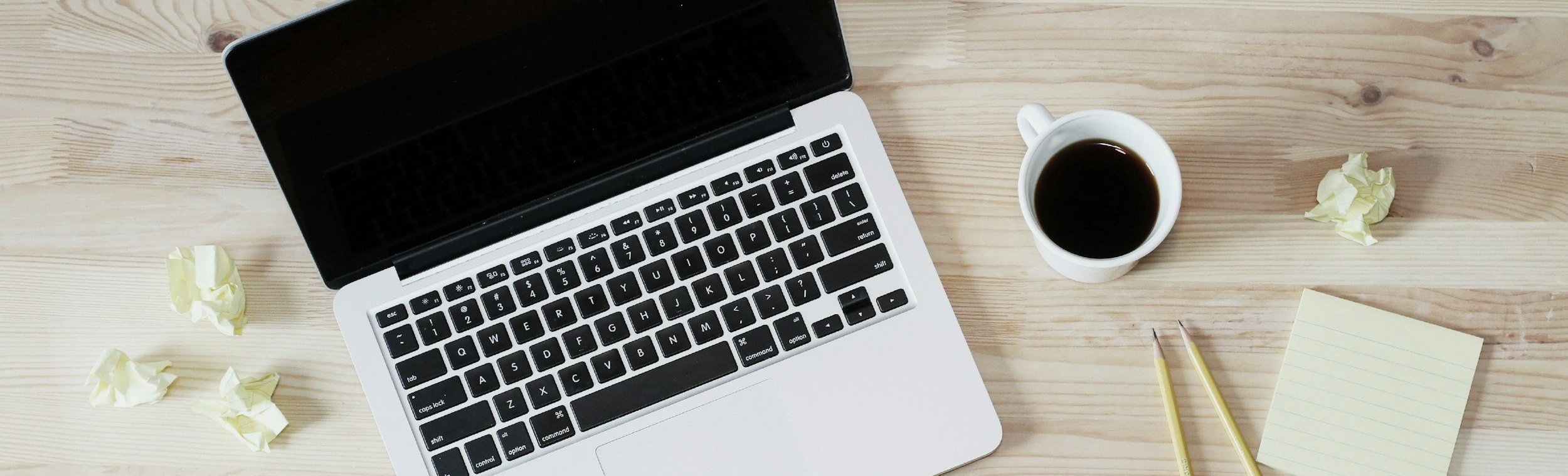 ノートパソコン、白いコーヒーカップのコーヒー、二本の黄色いペン、付箋紙、散らばった紙くずが木製の机の上に置かれている風景